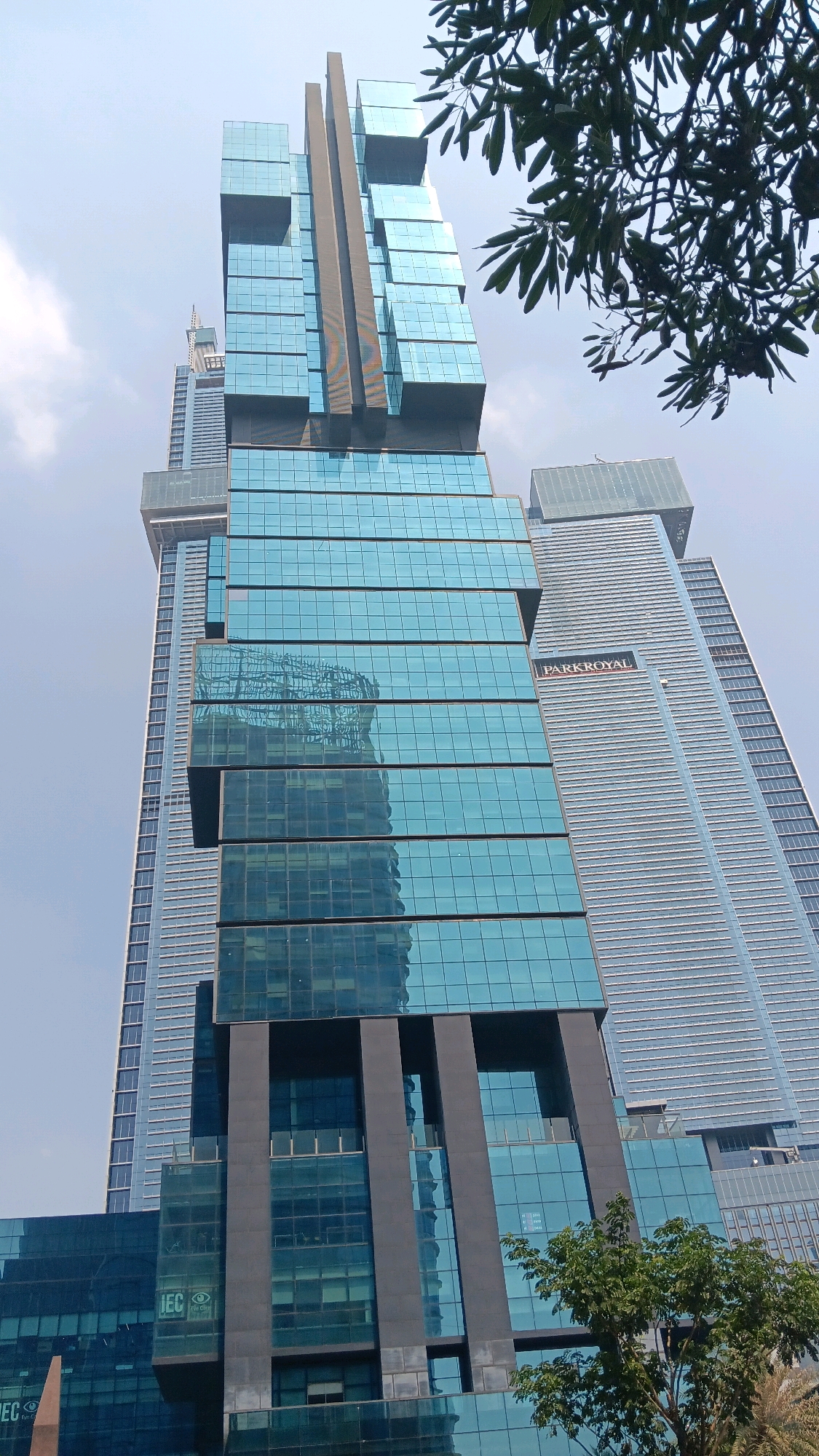 This image captures a low-angle view of the modern architecture of the Lumina building in Central Jakarta, Indonesia, on a clear day. The Lumina building, with its distinctive blue glass facade and stacked geometric design, dominates the frame. To its right, the PARKROYAL hotel is visible, its signage clearly displayed on its sleek, silver-toned exterior. In the foreground, lush green foliage from a tree partially obscures the lower sections of the Lumina building. Other skyscrapers, characteristic of a major metropolitan area like Central Jakarta, form the backdrop, contributing to the dense urban landscape. The sky is a pale blue with a few wispy clouds. No people are visible in the image.