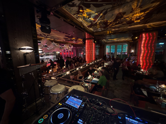 The image displays the interior of a dimly lit, high-end restaurant or bar located in the city of London, United Kingdom. In the immediate foreground, a professional DJ setup with illuminated screens and controls is visible on the right, accompanied by two conga drums positioned centrally. To the left, a dark vertical wall panel features a modern, horizontal sconce light.
The midground reveals a populated dining and lounge area. A long bar runs along the left side, stocked with bottles and glassware, and attended by at least one bartender. Overhead, multiple large, white, amorphous sculptural light fixtures are suspended. Numerous patrons, appearing to be adults, are seated at various tables and booths throughout the central and right sections of the venue, engaged in dining, drinking, and conversation.
Architectural details include several prominent, tall columns illuminated with a vibrant red light, dark paneled walls, and an ornate ceiling featuring gold and red patterns, accented by dynamic light projections. In the background, large, grid-patterned windows provide a blurred view of exterior lights, appearing green and blue. The overall environment is characterized by a dense gathering of individuals within a richly decorated space, featuring specialized lighting and entertainment equipment.