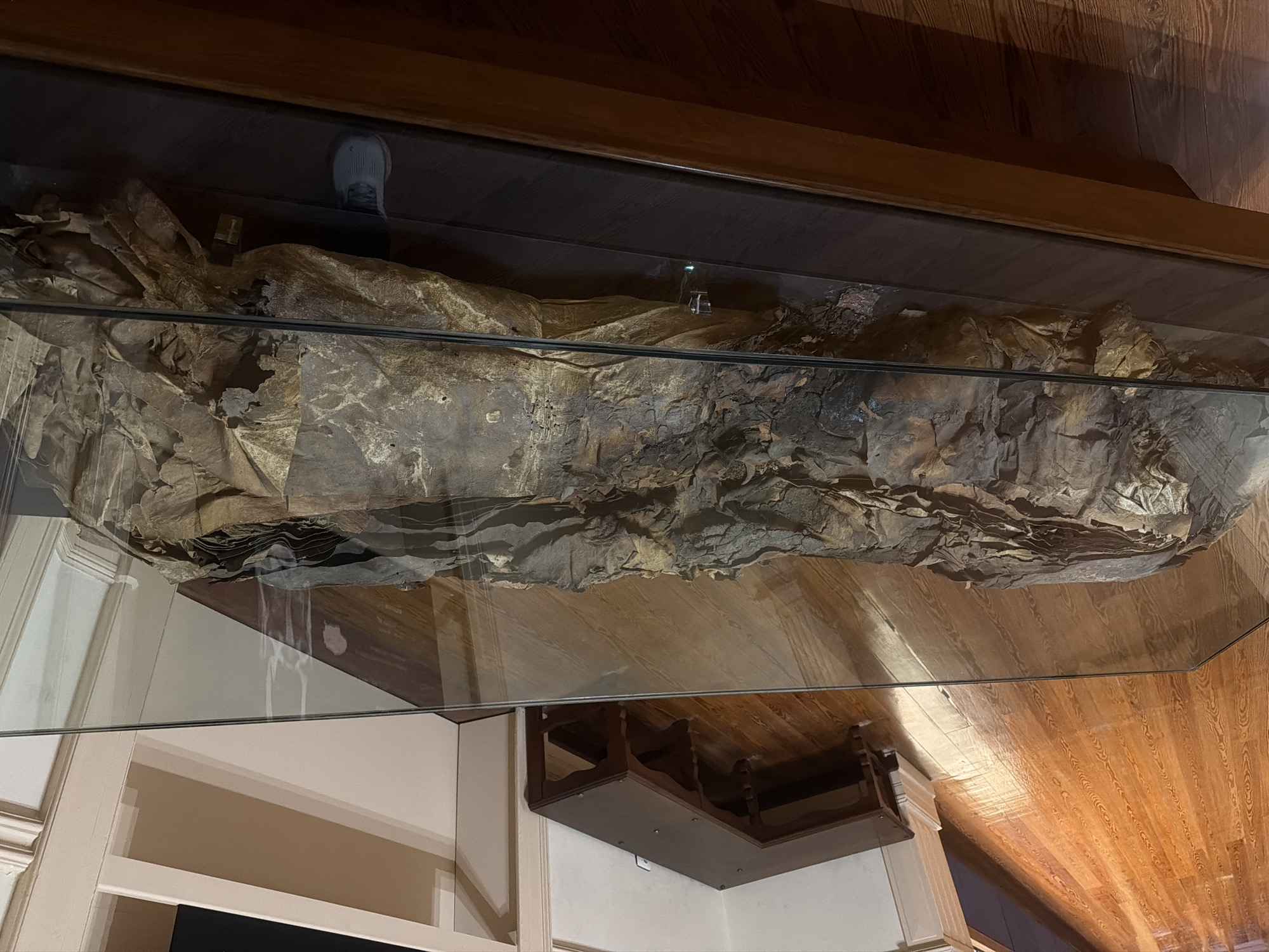 A preserved mummy is displayed in a glass-covered wooden case. The mummy is wrapped in brown, tattered cloth and appears to be lying on its back. The case is situated on a wooden floor with a rich, warm tone. In the background, a dark wooden L-shaped table with carved legs is visible, positioned against a white wall with architectural molding. To the right, a section of a white shelf unit can be seen. The overall scene suggests an indoor exhibition, possibly a museum or historical exhibit. The specific location in Las Palmas De Gran Canaria, Spain, is not determinable from the visual information provided in the image.