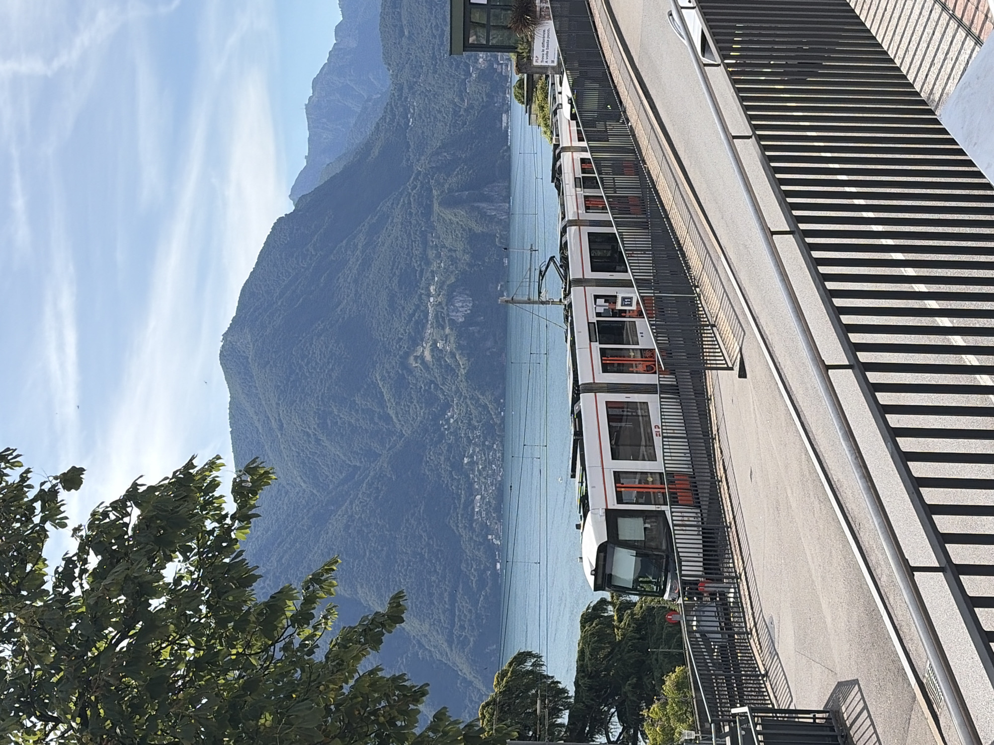 The image shows a modern, white and orange tram on a track that curves to the right, ascending an incline. The tram has multiple carriages and large windows. In the background, a large, bright blue lake stretches out, bordered by lush, green mountains under a clear blue sky with wispy clouds. The mountains are covered in trees and dotted with buildings, suggesting a populated area. In the foreground, a dark metal railing runs along the edge of a paved pathway. The overall scene depicts a scenic view in Lugano, Switzerland, likely from an elevated viewpoint overlooking the lake and the city.
