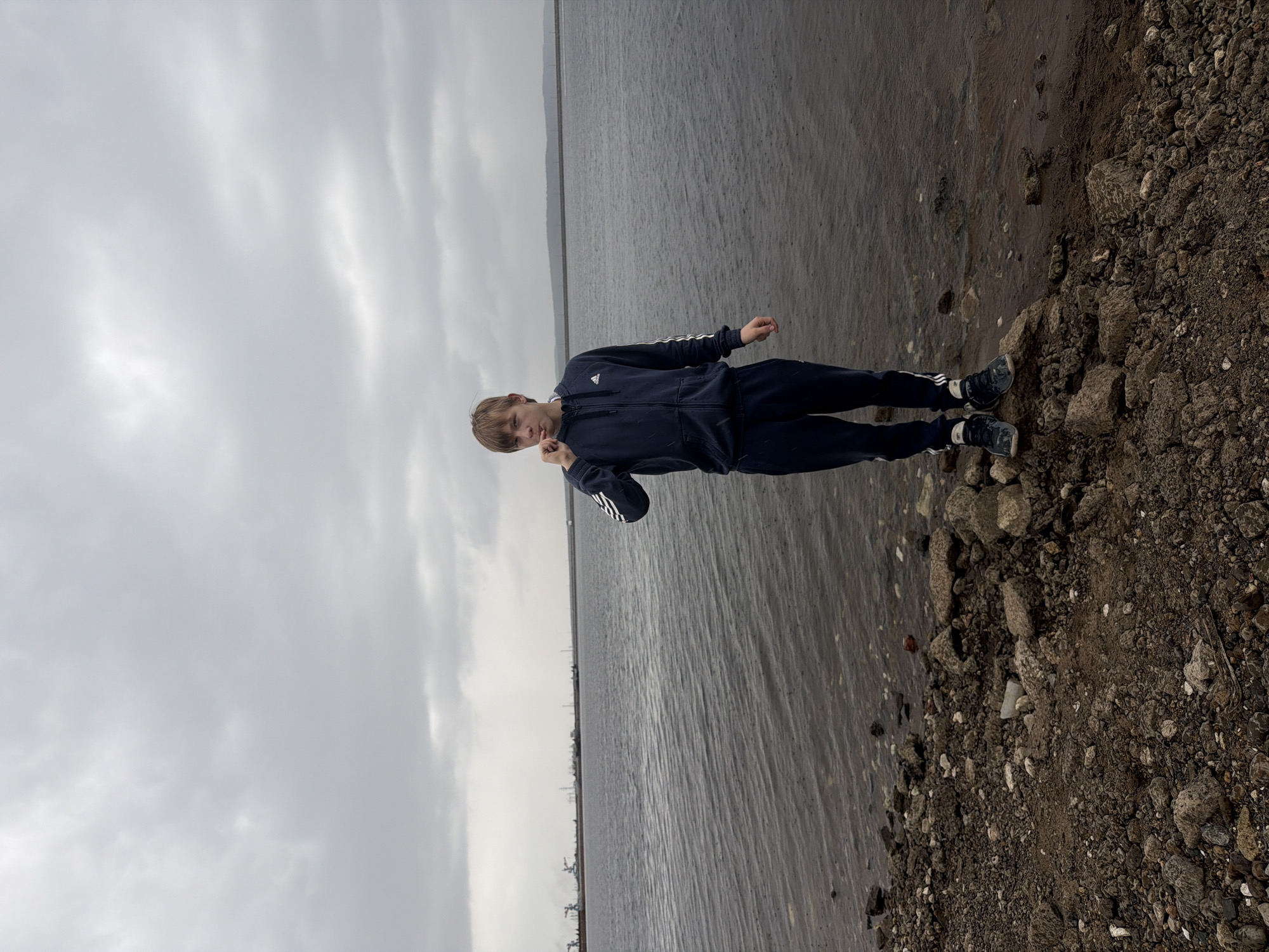 A young person stands on a rocky embankment near a body of water under a grey sky. The person wears a dark tracksuit with white stripes. The body of water is flat with industrial structures visible in the distance. The embankment is made of small rocks and debris. The image was taken near the city of Chaikovsky, Russia.