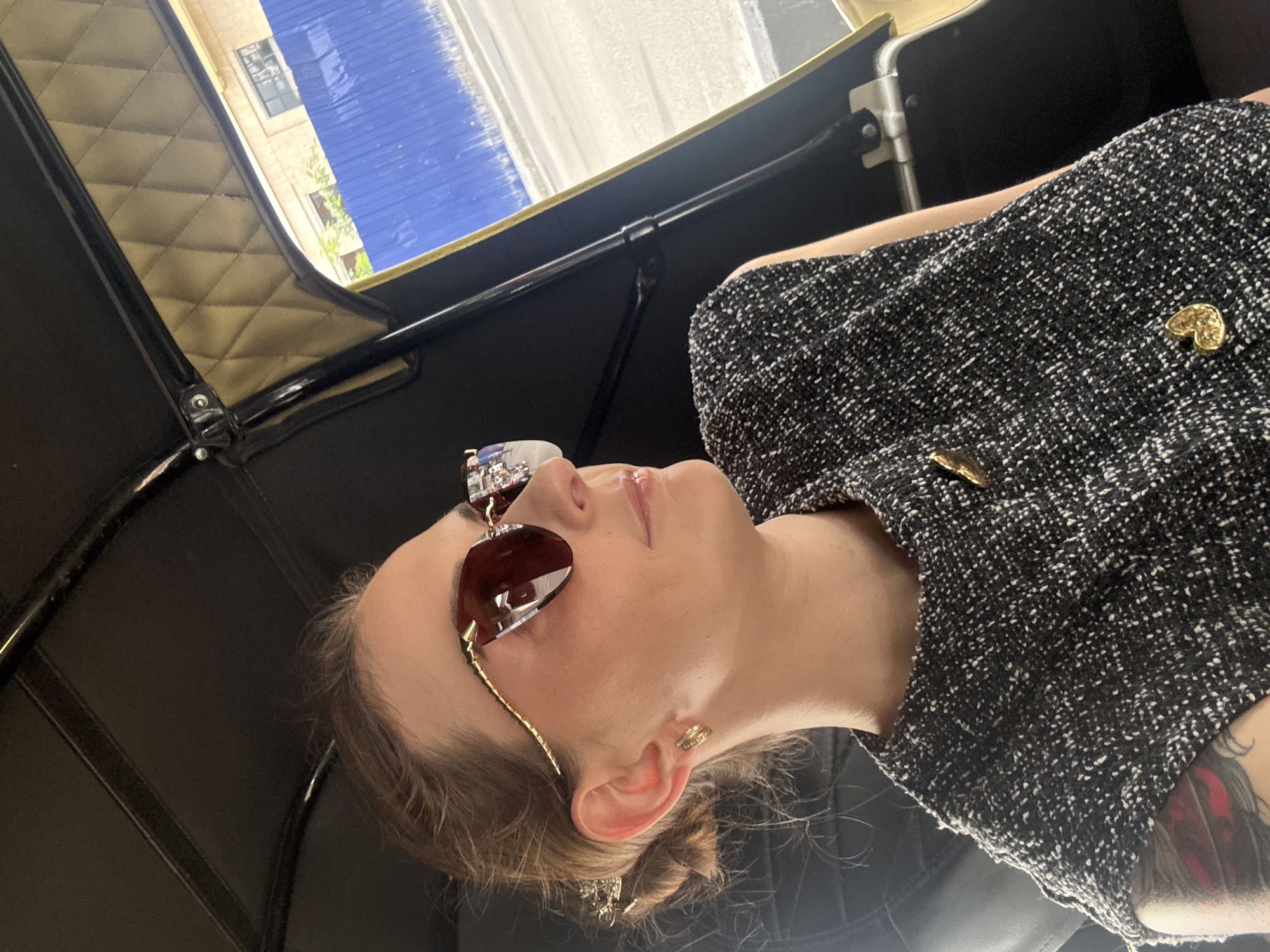 A medium shot photograph, taken indoors with an upward angle, shows a woman seated inside a vehicle, likely a tuk-tuk, with the exterior view visible through an opening. The woman, who is the primary subject, is facing right, with her head slightly turned upwards. She has her hair pulled back into a bun and is wearing dark sunglasses with a gold-colored frame that has a pointed design extending from the temples. She also has on a sleeveless, tweed-like top with a speckled black and white pattern, adorned with two gold heart-shaped brooches. A delicate gold chain necklace with a pendant is visible around her neck. Her left earlobe features a small, gold hoop earring. On her left arm, a tattoo with red and black ink is partially visible. The interior of the vehicle features dark upholstery and a quilted, golden-tan headliner. Through the opening of the vehicle, a bright blue construction fence is prominent, suggesting an urban environment undergoing development. Behind the fence, a building with windows is visible, and the ground appears to be a paved road or sidewalk. The overall impression is of a woman traveling in a city, possibly during the daytime, given the lighting and the reflection in her sunglasses which shows a brightly lit outdoor scene with possibly other vehicles. The presence of a tuk-tuk is a common sight in cities like Phnom Penh, Cambodia, where Khan Prampir Makara is a district.