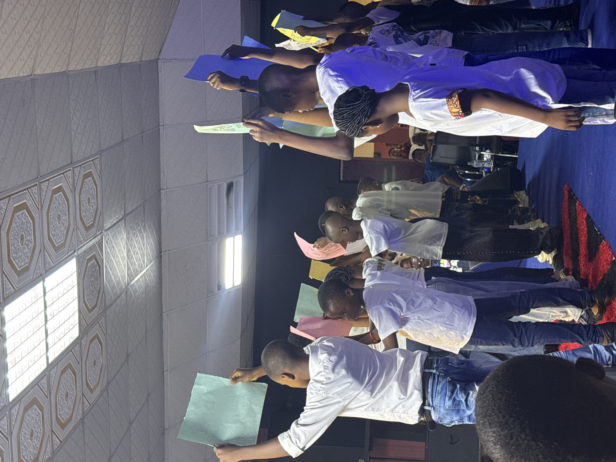 A group of youths dressed in white shirts and jeans are holding up colored paper in a brightly lit room in Nkpor, Nigeria. Some are reading from the paper. The room has patterned tile walls and a blue-carpeted area.