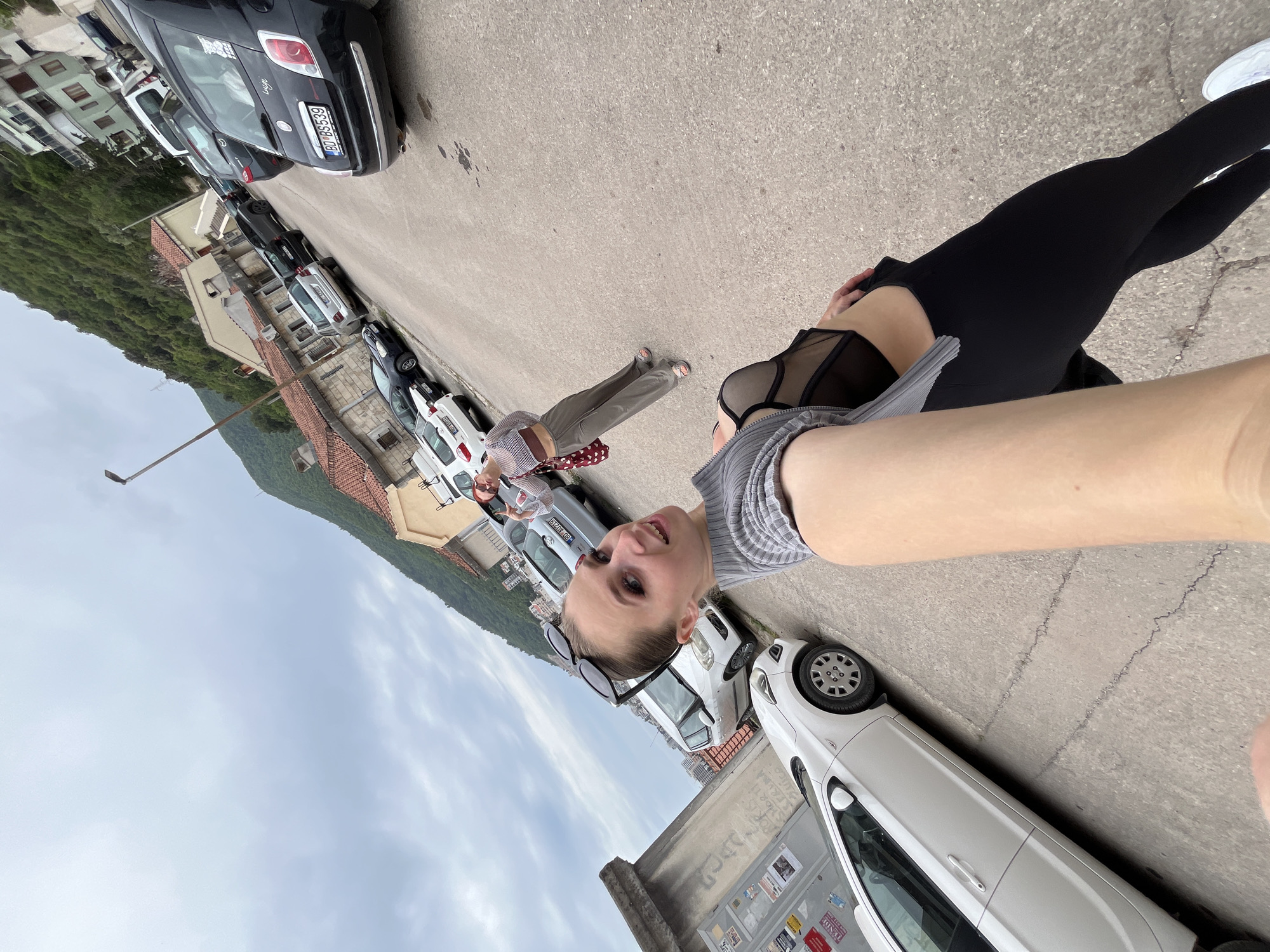 The image is an eye-level self-portrait of a young woman standing on a street in Budva, Montenegro. She is wearing sunglasses on her head, a gray top, black leggings, and white sneakers. The woman is positioned close to a row of parked cars. In the background, another person is visible further down the street. The street is flanked by buildings on one side and a mountainside covered in trees on the other. The sky is overcast with visible clouds. Cars of various colors are parked along the street. The setting is a narrow road with typical European architecture. A street lamp is also visible along the road.