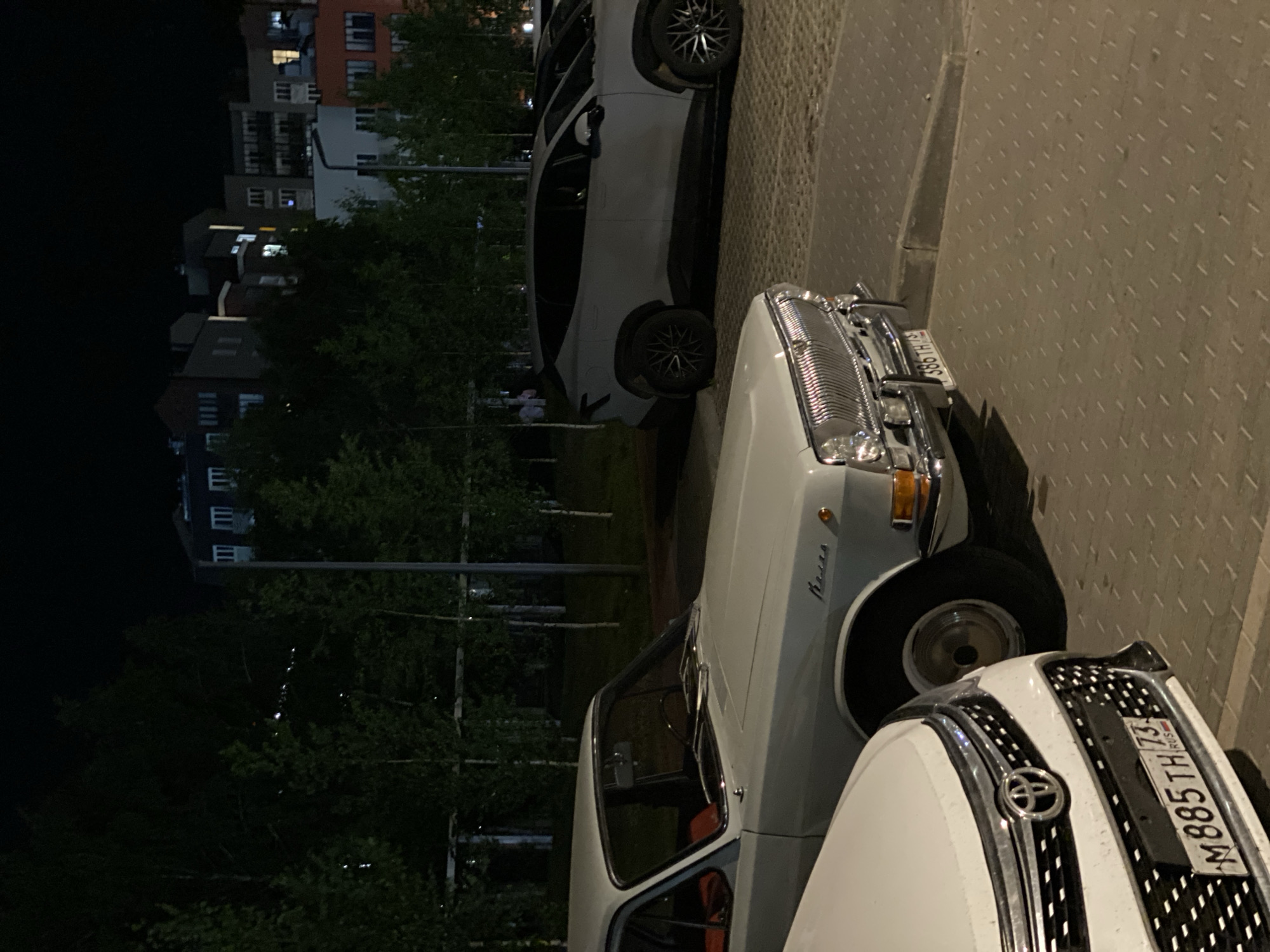 The image captures a nighttime scene in Ulyanovsk, Russia, featuring several cars parked in a paved lot adjacent to a wooded area and apartment buildings. In the foreground, the front of a white Toyota is visible, displaying a license plate that reads "M885TH 73 RUS." To its right is a vintage white car, possibly a GAZ, with a chrome grille and a license plate reading "386TH 73." Further in the background, a modern white SUV is parked, and beyond that, a row of multi-story apartment buildings is illuminated against the dark sky. The trees in the middle ground are dense and dark, with some lights visible through them, suggesting the proximity of the buildings. The paved ground is textured with a pattern of raised dashes. The overall atmosphere is quiet and urban, characteristic of a residential area at night.
