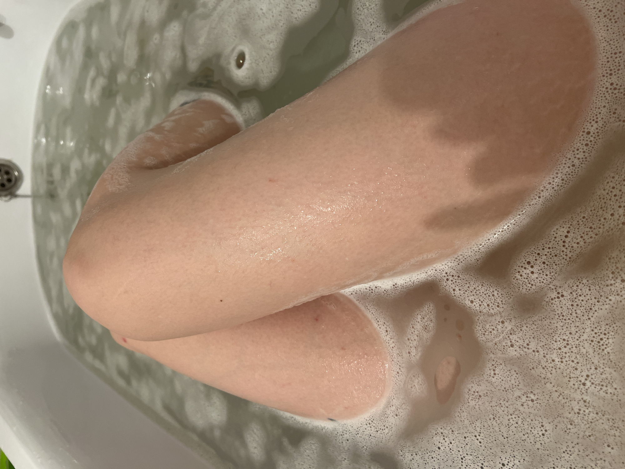 A top-down, high-angle close-up shot shows a portion of a person's legs and feet in a bathtub filled with soapy water. The person's legs are crossed, with the top leg partially submerged and the bottom leg mostly underwater. The water is bubbly and foamy, obscuring the bottom of the tub. A drain is visible in the upper-left corner of the frame. The location is not discernable from the visual information provided.