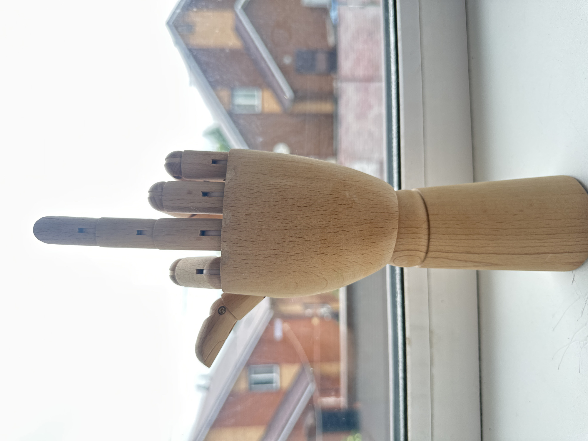 A wooden mannequin hand is positioned upright on a white windowsill. The hand is held in a gesture where the middle finger is extended, while the thumb and other fingers are closed. Behind the hand, through a window, the facade of a two-story brick house with a gabled roof is visible. The sky is overcast.