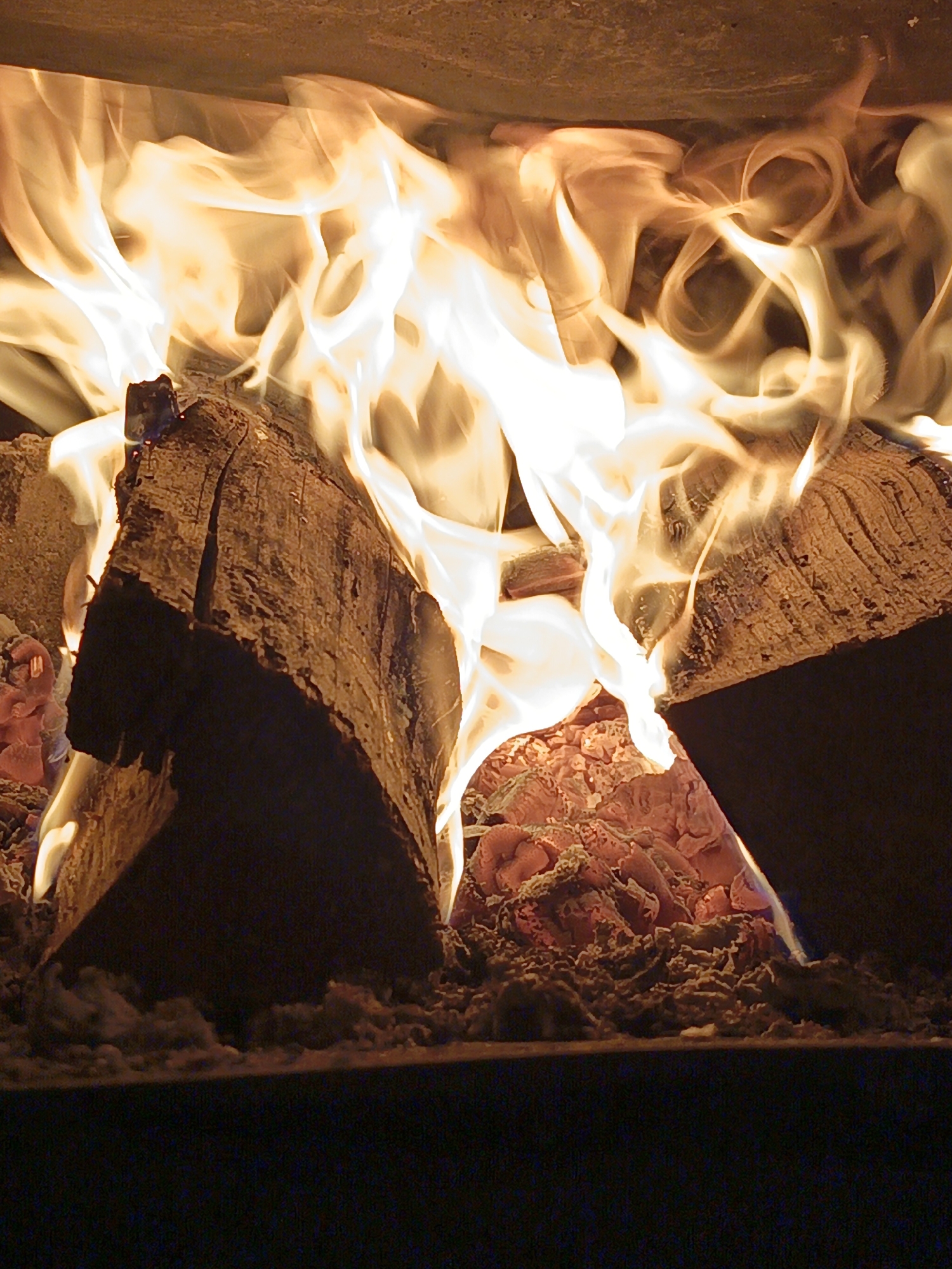 The image presents a close-up, vertical perspective of an active wood fire contained within an enclosure, likely a fireplace or firebox. Two primary logs, split and partially consumed, are centrally positioned, forming a gap between them. Intense, bright yellow-orange and white flames rise vigorously from these logs and the substantial bed of glowing red and orange embers and ash beneath them, illuminating the scene. The flames exhibit a fluid, somewhat blurred appearance due to motion. The logs display visible wood grain and charring. The upper portion of the frame reveals a warm-toned, textured surface, likely the interior wall or ceiling of the fire enclosure, brightly lit by the flames. The bottom portion of the image is dark and indistinct, suggesting the floor or opening of the fire chamber. No individuals or specific identifying markers for a geographical location, such as Лосева, Belarus, are discernible within the image.