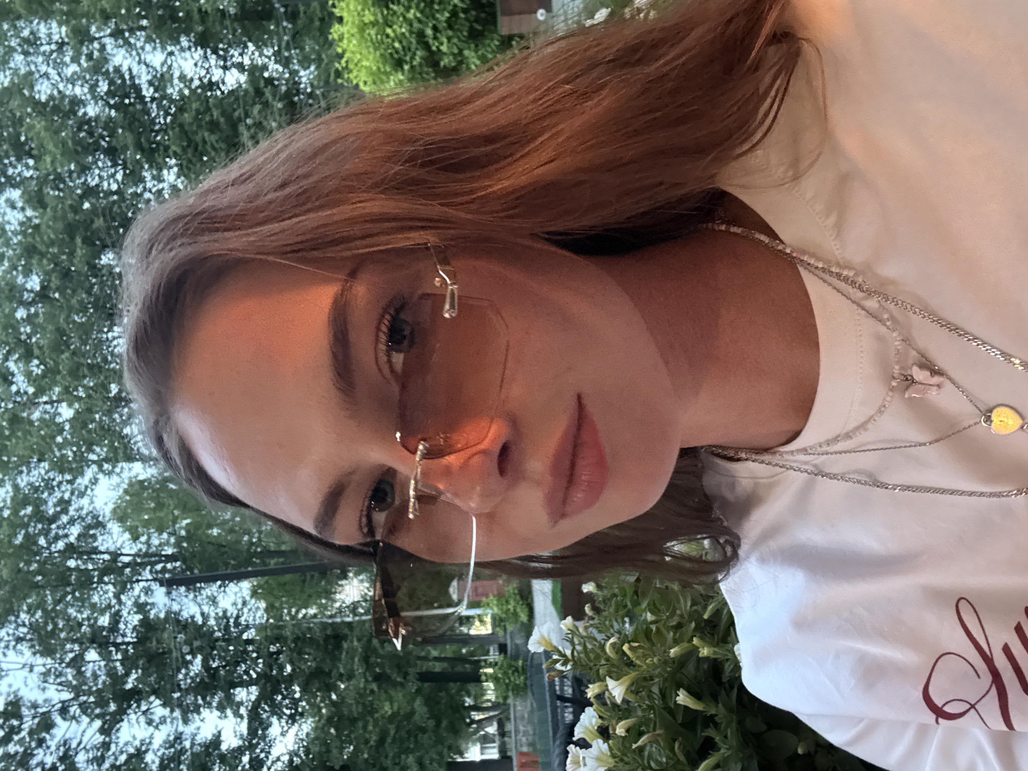 A young adult female is captured in a close-up outdoor portrait, looking directly at the viewer. She has long, auburn-brown hair, parted in the middle and flowing over her shoulders. She wears rimless, amber-tinted sunglasses with minimal gold-toned metal detailing, through which her blue-grey eyes are visible. Her nose bridge is prominent, and her lips are full and slightly parted, displaying a neutral expression. A strong, warm golden light illuminates the right side of her face, hair, and shoulder. She is dressed in a plain white crew-neck t-shirt, with a partial red/maroon script or design visible on the lower left of the garment. She accessorizes with layered necklaces: a short, light-colored beaded necklace featuring a pink butterfly charm, a thin silver-toned chain, and a longer silver-toned chain ending in a small, possibly gold-toned, heart-shaped locket. To her immediate left in the foreground, a bush with numerous white, trumpet-shaped flowers is in bloom. The background consists of dense green foliage from tall trees and bushes, with glimpses of a lighter sky and indistinct urban elements like distant buildings and a dark pole visible through the trees. The scene is located in the city of Perm, Russia.
