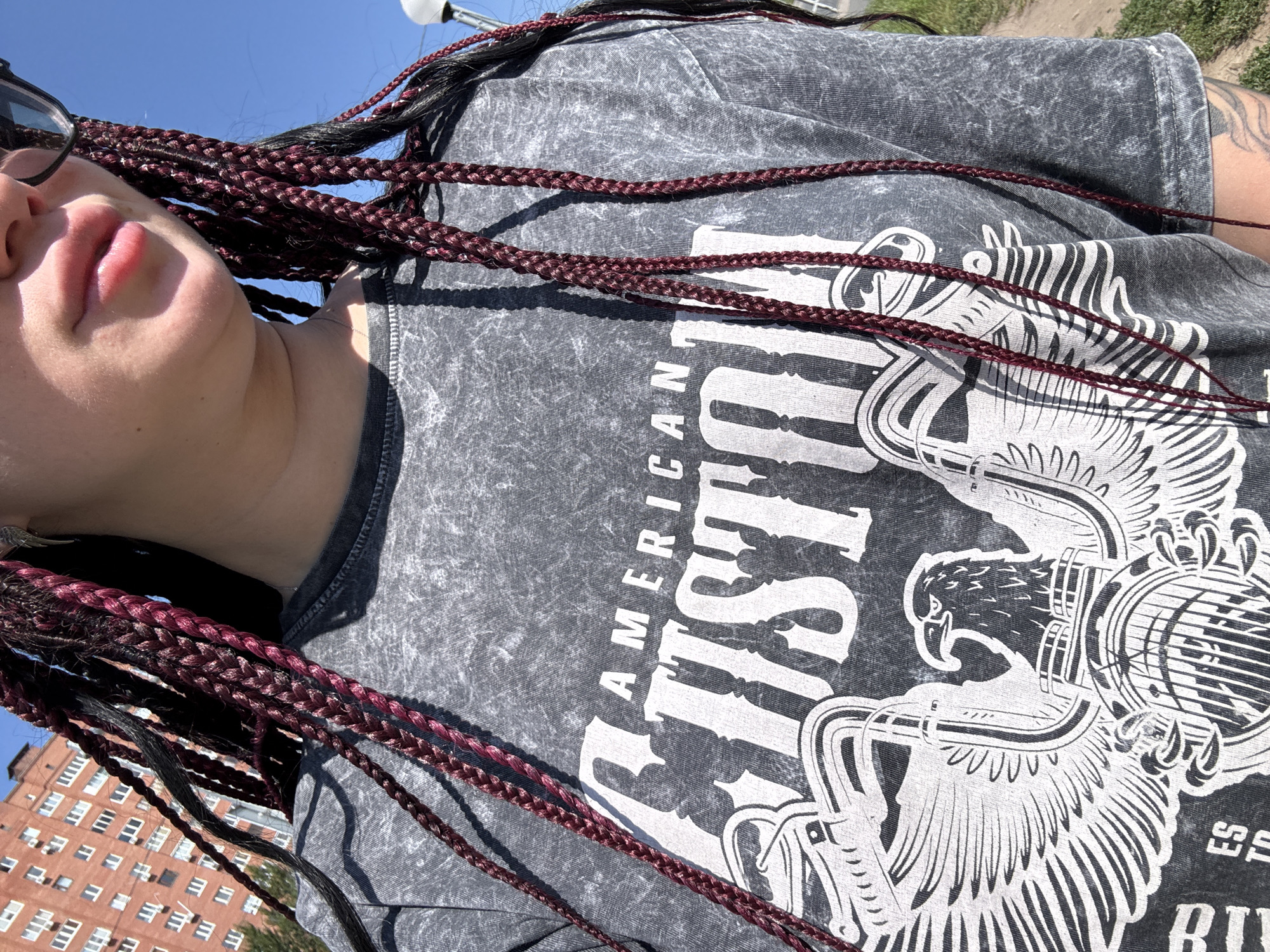 A person stands outdoors on a sunny day in Tolyatti, Russia, wearing a grey t-shirt with a graphic print. The t-shirt features large white lettering that reads "AMERICAN CUSTOM" in a stylized font, layered over an illustration of an eagle with outstretched wings. The person's hair is styled in numerous braids, some of which are dark and others have a burgundy hue, and these braids hang down over the t-shirt. In the background, a multi-story brick building with many windows is visible. The person's lower face and lips are partially visible, and they appear to be wearing sunglasses on their head, though they are not currently on their face. The environment suggests an urban setting, possibly a residential area, with a paved area or sidewalk visible to the right of the person and some greenery.