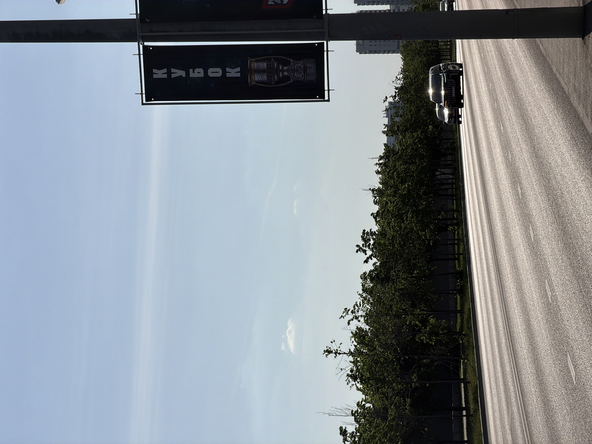 The image depicts a street scene in Kazan, Russia. A black banner with white Cyrillic text ("KABOK") and an image of a trophy hangs from a lamppost. A roadway runs parallel to a line of trees. Two cars are visible on the road. The sky is light blue with a few wispy clouds. Buildings are partially visible in the background. The lighting suggests it is daytime, possibly late afternoon or early morning. The image is oriented vertically.