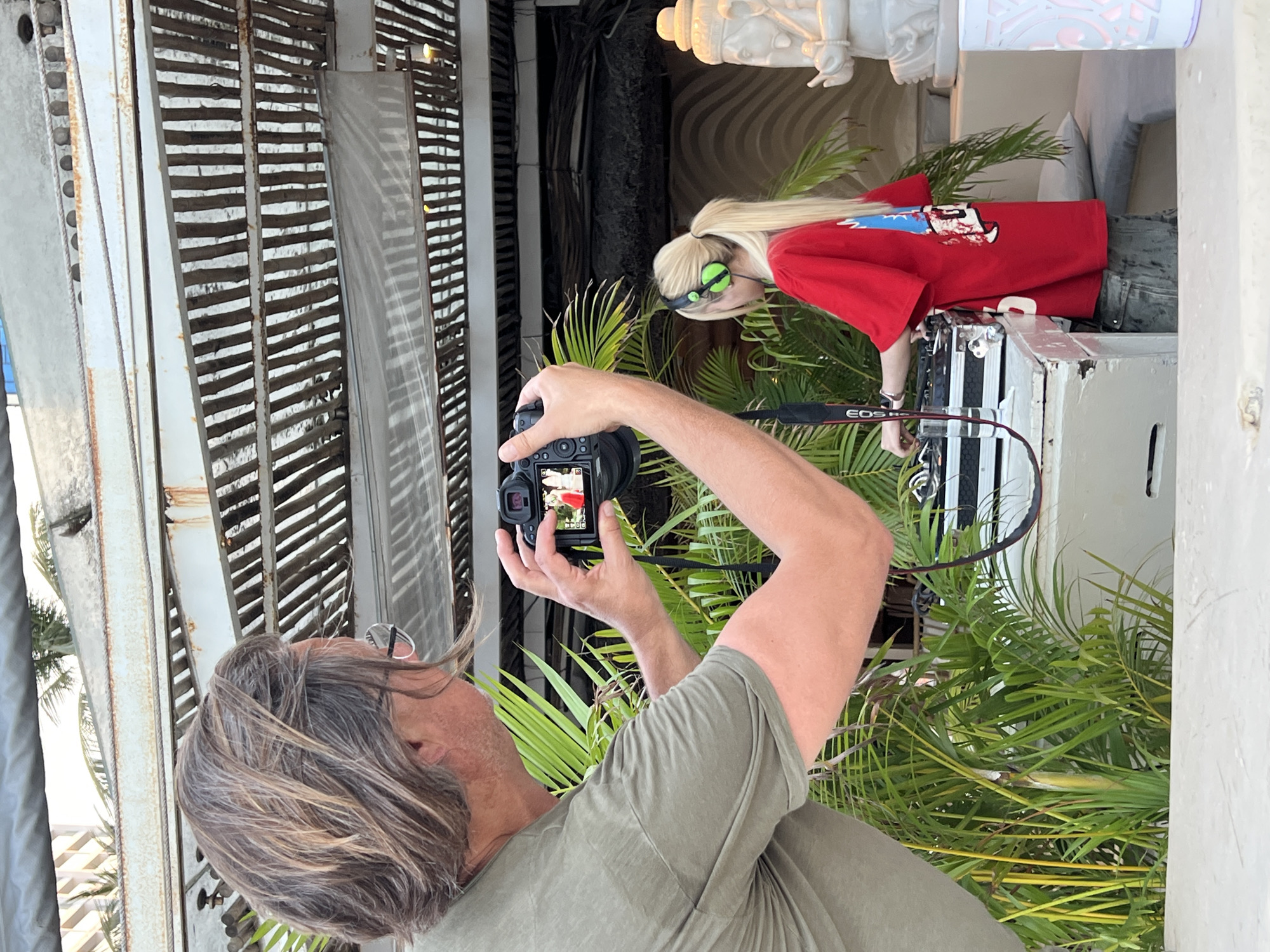 A man photographs a blonde woman in Nha Trang, Vietnam. The man, with graying hair and glasses, wears an olive-colored t-shirt and holds a DSLR camera with a red flower visible on the screen. The woman has a blonde ponytail and wears green headphones and a red shirt. She stands near a white object, potentially a speaker, surrounded by tropical foliage. The backdrop features gray metal slats and glimpses of a white column or statue.