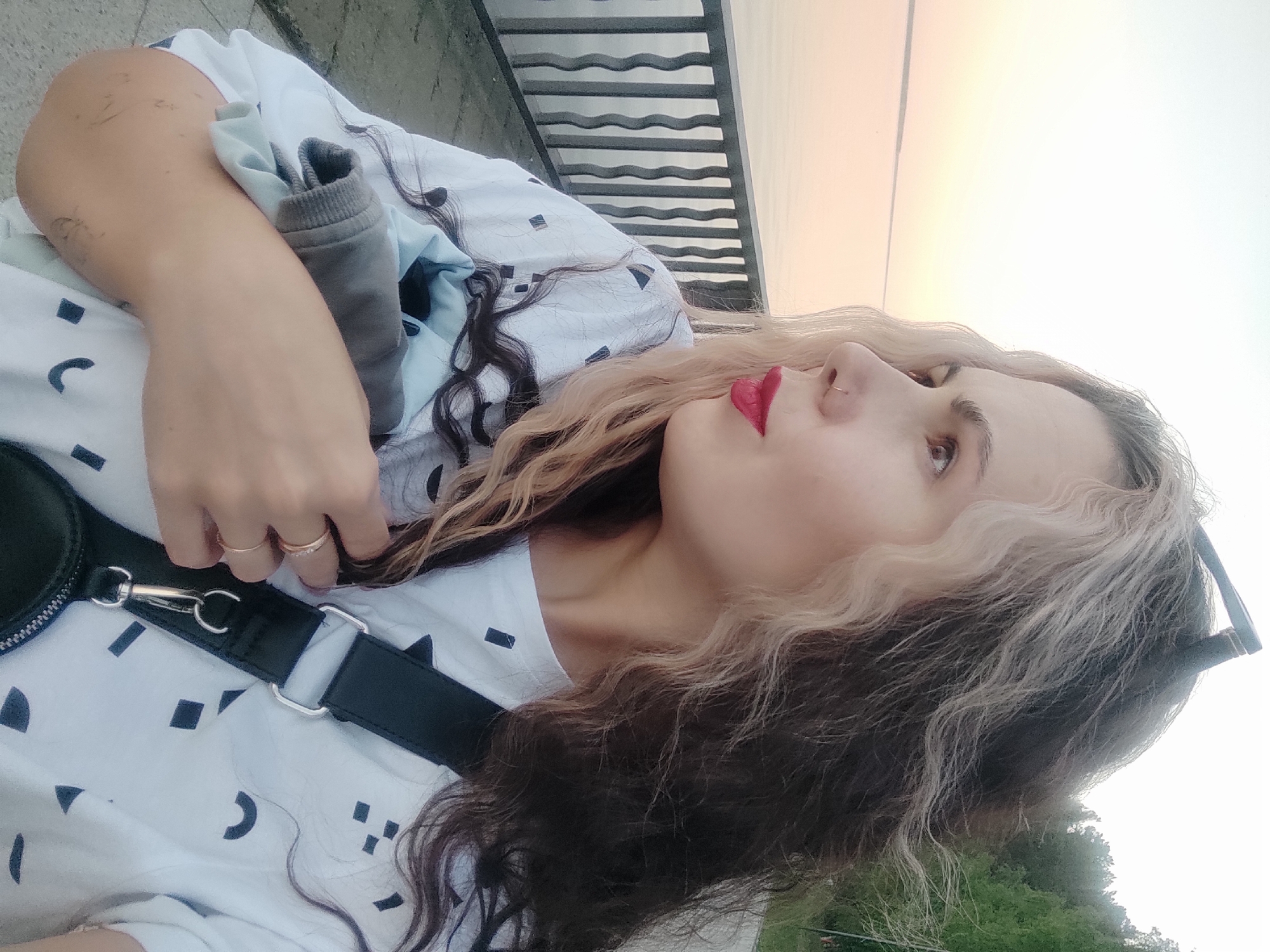 A young woman with dark and light blonde streaked hair, wearing red lipstick and two gold rings, is seated outdoors in an unknown city. She is dressed in a patterned white t-shirt with a black shoulder strap attached to a black bag. She holds a light blue sweater with a gray sleeve in her right hand. Behind her is a metal railing with a body of water and a hazy sunset in the background. There is some green vegetation visible in the lower right corner of the image.