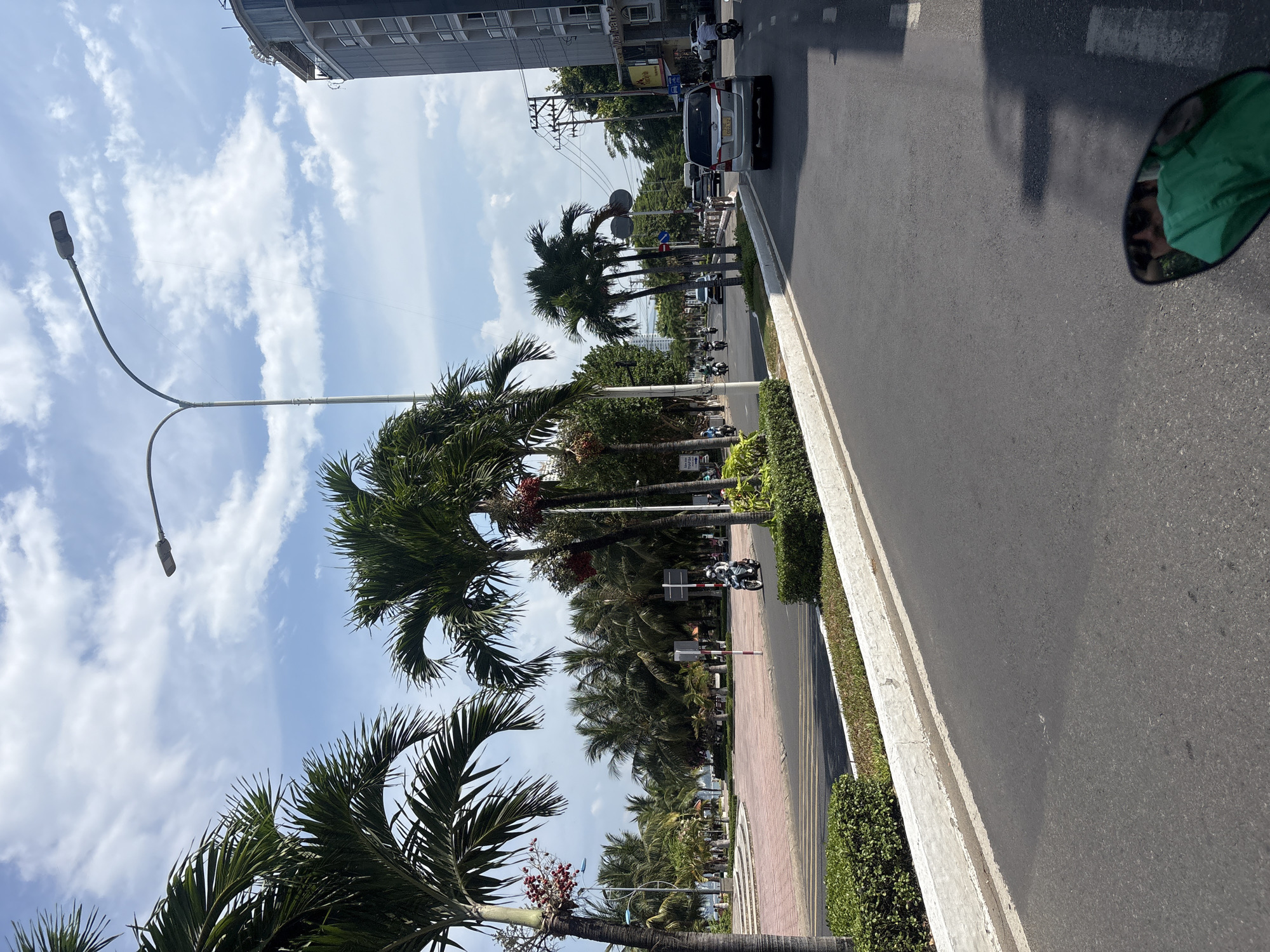 This image depicts a street scene in Nha Trang, Vietnam, likely along the coastal road. The perspective is from someone on a motorcycle, as evidenced by the motorcycle's mirror reflecting the rider's face and a green garment. The street is paved with asphalt and lined with manicured hedges and palm trees. A multi-story modern building with balconies is visible on the right side of the road, suggesting an urban or resort area. In the background, more palm trees and some smaller buildings are present. Several motorcycles and a silver car are in motion on the road. The sky is partly cloudy with a bright, daytime sun. The overall impression is a sunny day in a tropical city.