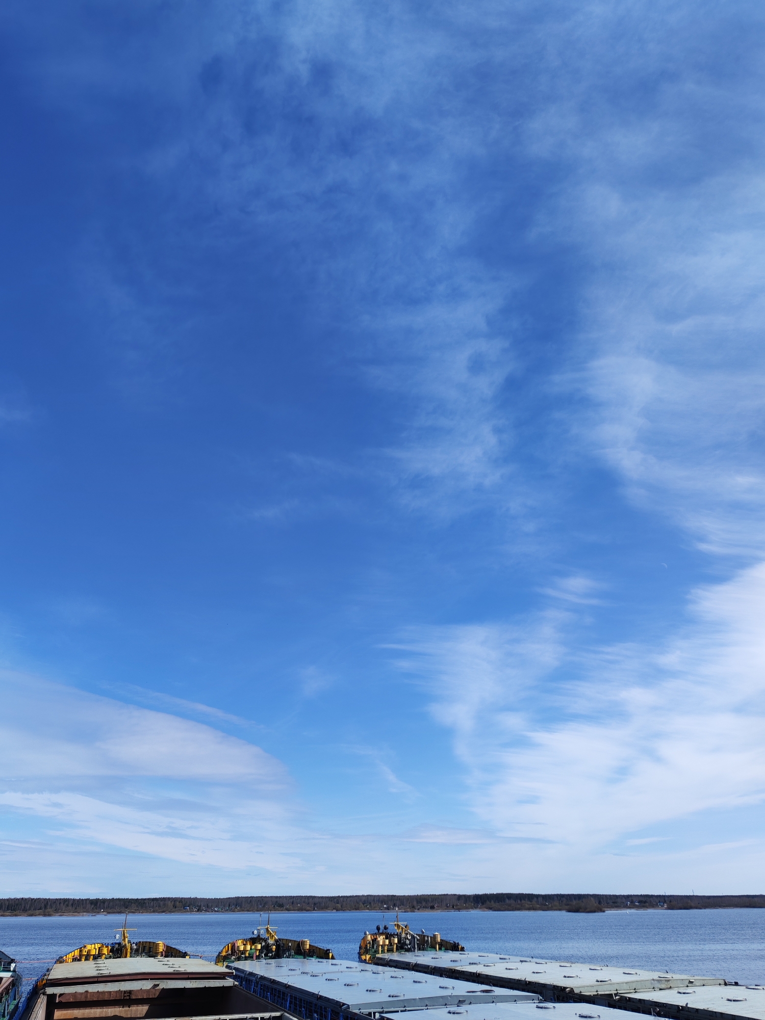 The image captures a bright, sunny day in Rybinsk, Russia, with a wide expanse of blue sky partially filled with thin, wispy clouds. The lower portion of the frame shows a body of water, likely the Volga River, with a distant shoreline visible. A line of trees or a forest borders the water's edge. Several flat-deck barges are in the foreground, likely docked or stationary on the river. These barges appear to be carrying some form of equipment or materials, indicated by the yellow-colored structures on top.
