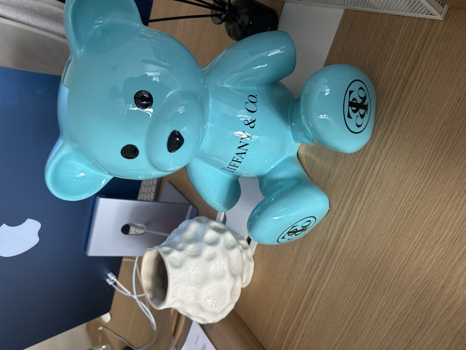 This scene depicts an indoor desktop setting, situated in Paris, France. A prominent, glossy, light blue ceramic teddy bear figurine, functioning as a piggy bank with a visible coin slot on its head, occupies the central foreground. The teddy bear has black eyes and a black nose, with "TIFFANY & Co." printed in black on its chest and the stylized "T&Co" monogram in black on each of its feet. To the left of the teddy bear, a white, irregularly textured ceramic vase or pot with a wide opening is positioned. Behind these items, a dark blue Apple iMac computer monitor is visible, displaying a silver Apple logo on its rear surface. A silver computer stand or dock sits directly in front of the iMac's base. White electrical cables are discernible behind the vase and near the computer setup. On the far right, a dark-colored bottle containing multiple black diffuser reeds is partially visible. The objects rest upon a light-colored wooden desk surface characterized by horizontal grain patterns. A piece of white paper is partially visible beneath the white vase, and a portion of a white mesh accessory is present at the far right edge of the desk. No people are present, and no active interactions are occurring.