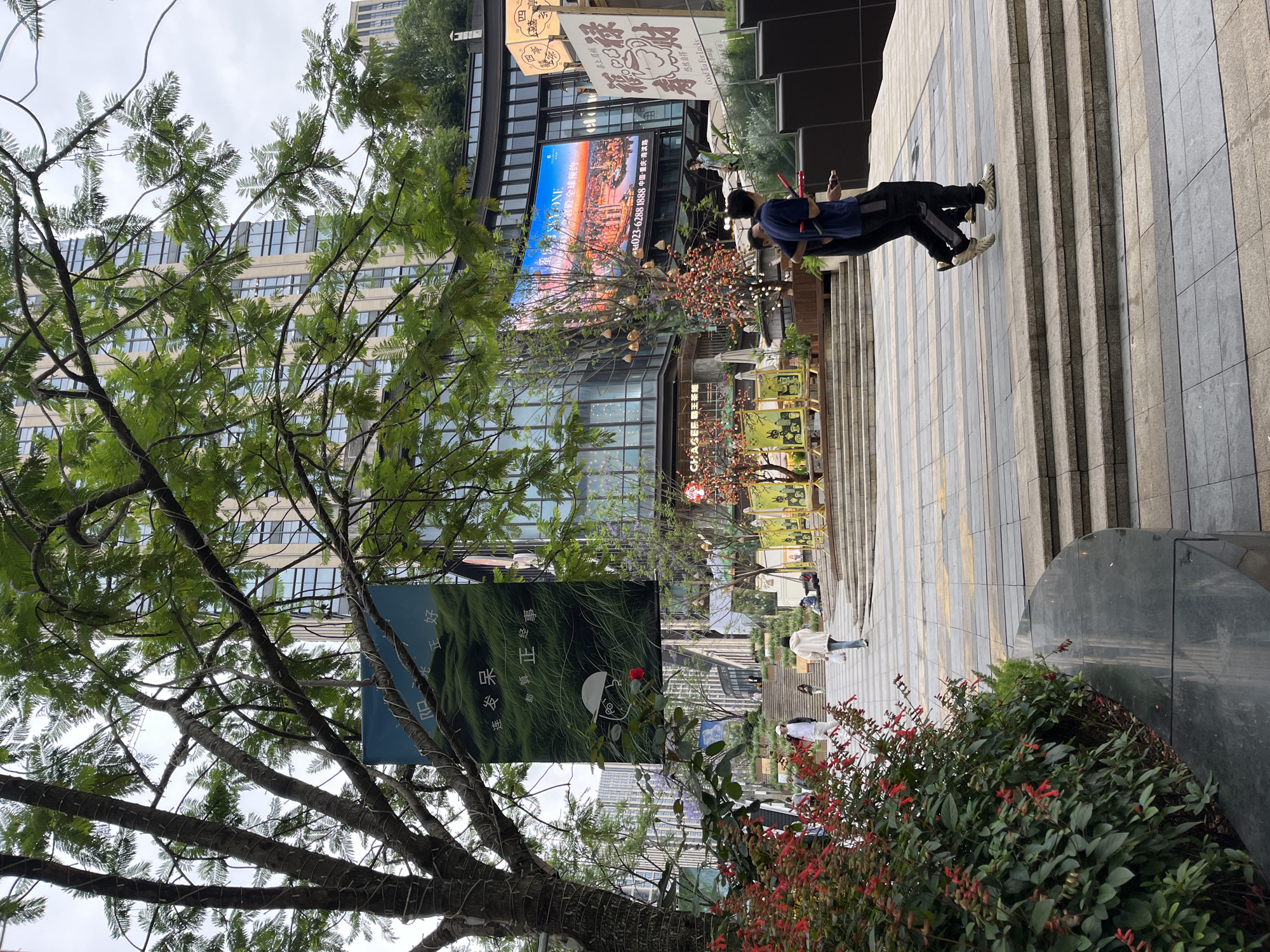 The image shows a street scene in Yuzhong District, China. The scene includes a pedestrian area with steps, plants and several people walking. There is a tree on the left side of the image. In the background are buildings, including one with a large LED screen and several advertising signs.