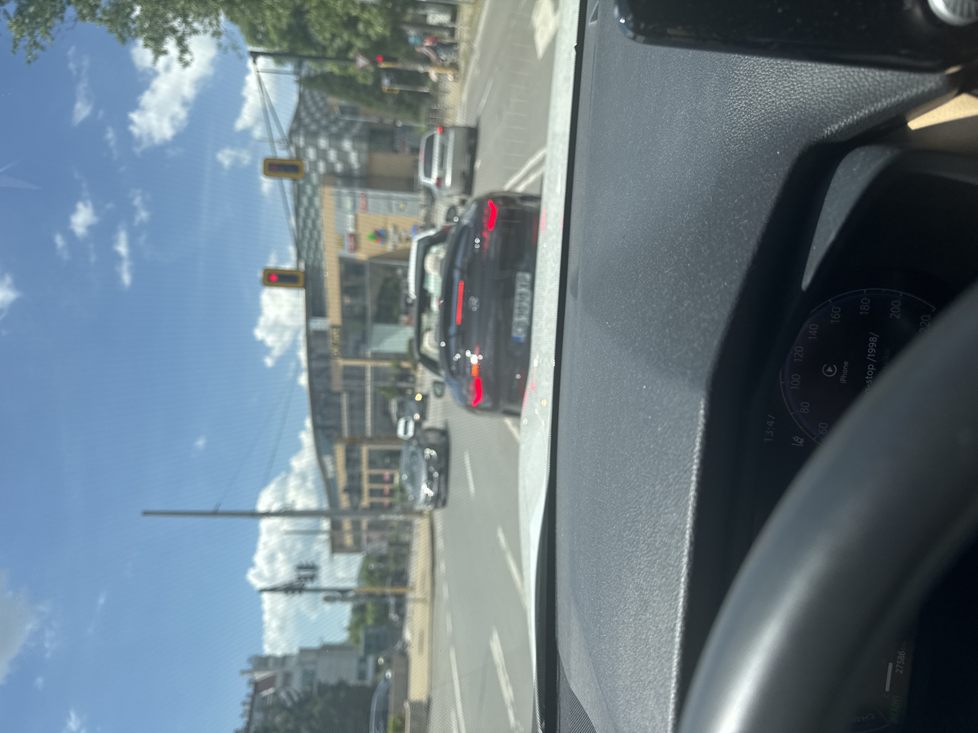 The image is taken from inside a car, showing the dashboard and steering wheel. Through the windshield, a street scene is visible, with several cars in front of the car, lined up at a traffic light which is red. Buildings and greenery are seen on either side of the road. Based on the license plate on the car ahead (containing "София"), this scene is in Sofia, Bulgaria. The car's odometer reads 27,586 km, and the time displayed on the dashboard is 13:47.