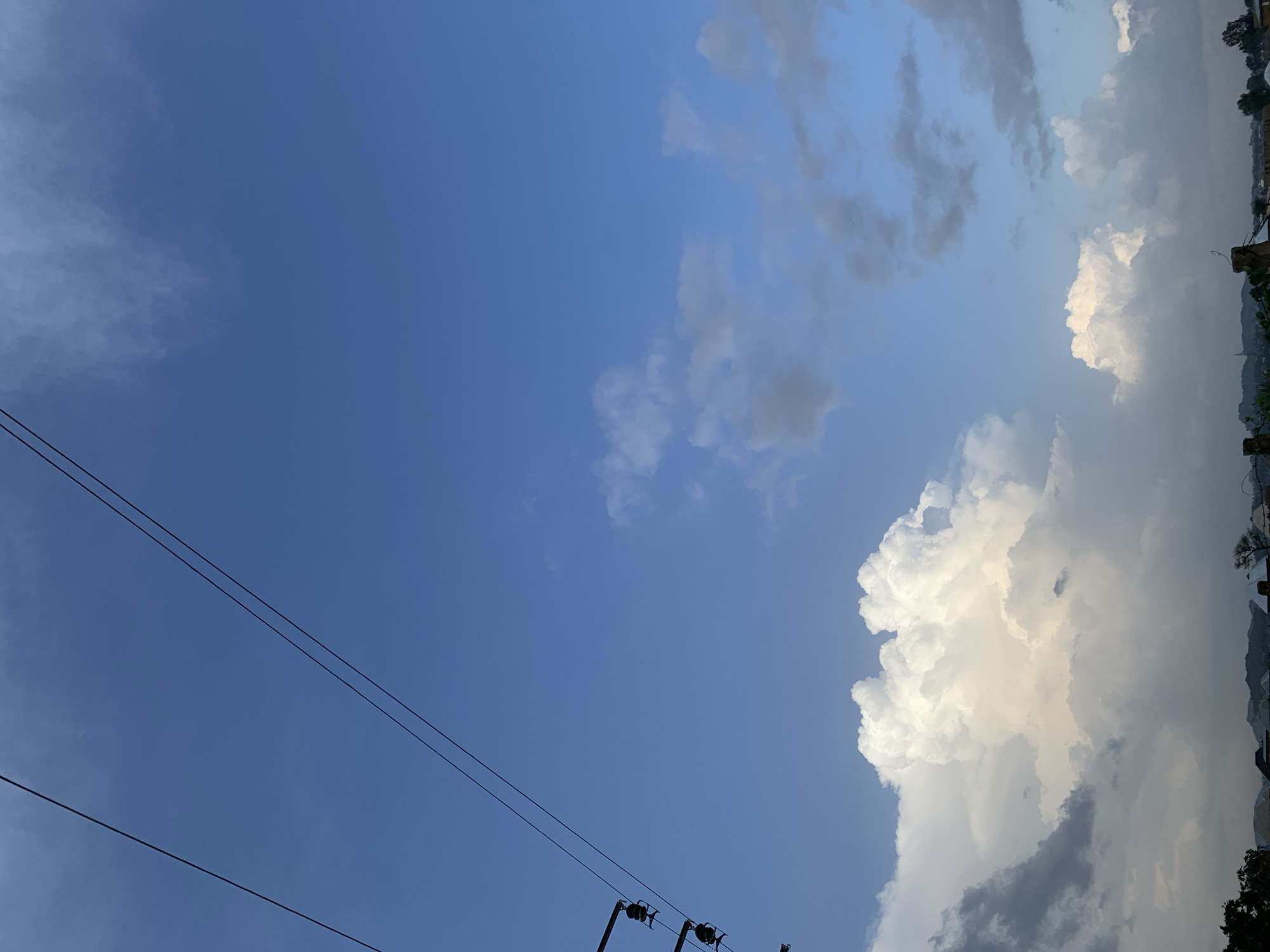 The image depicts a sky filled with a mix of blue sky and clouds, viewed from a low angle. In the foreground, two parallel black power lines stretch diagonally across the lower left portion of the frame, indicating an outdoor setting likely in a populated area. These lines are thin and taut, suggesting they are for electricity transmission. Further to the right, other power line components are visible, including what appear to be insulators and poles, hinting at infrastructure.
The sky is predominantly a clear, bright blue, with some wispy white clouds scattered in the upper left. The right side of the image features larger, more dramatic clouds. A large, bright white cumulus cloud dominates the lower right, illuminated from above and appearing almost luminescent. To its left and above, darker, grayer clouds suggest the presence of rain or storm clouds. This contrast between bright sunlight and shadowed clouds indicates a dynamic weather pattern, possibly a transition from clear to stormy weather or vice versa.
Along the far right edge of the image, a faint outline of land is visible, with indications of trees and possibly buildings or structures, suggesting a landscape or urban periphery. The overall impression is that of an open sky, possibly during the late afternoon or early evening, given the quality of light on the clouds. There are no people visible in the image. The mood is serene yet potentially dramatic due to the cloud formations. There is no visible text.