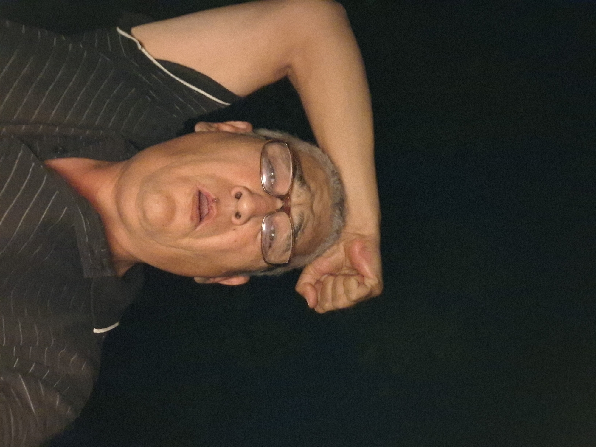 The image is a close-up, low-angle selfie of a middle-aged man with glasses, his right arm raised and fist clenched above his head. He is wearing a dark, pinstriped shirt. The background is dark, suggesting it is night or an indoor setting with minimal lighting. There is no discernible information in the image that would allow for the determination of the location within the city of Almaty, Kazakhstan.