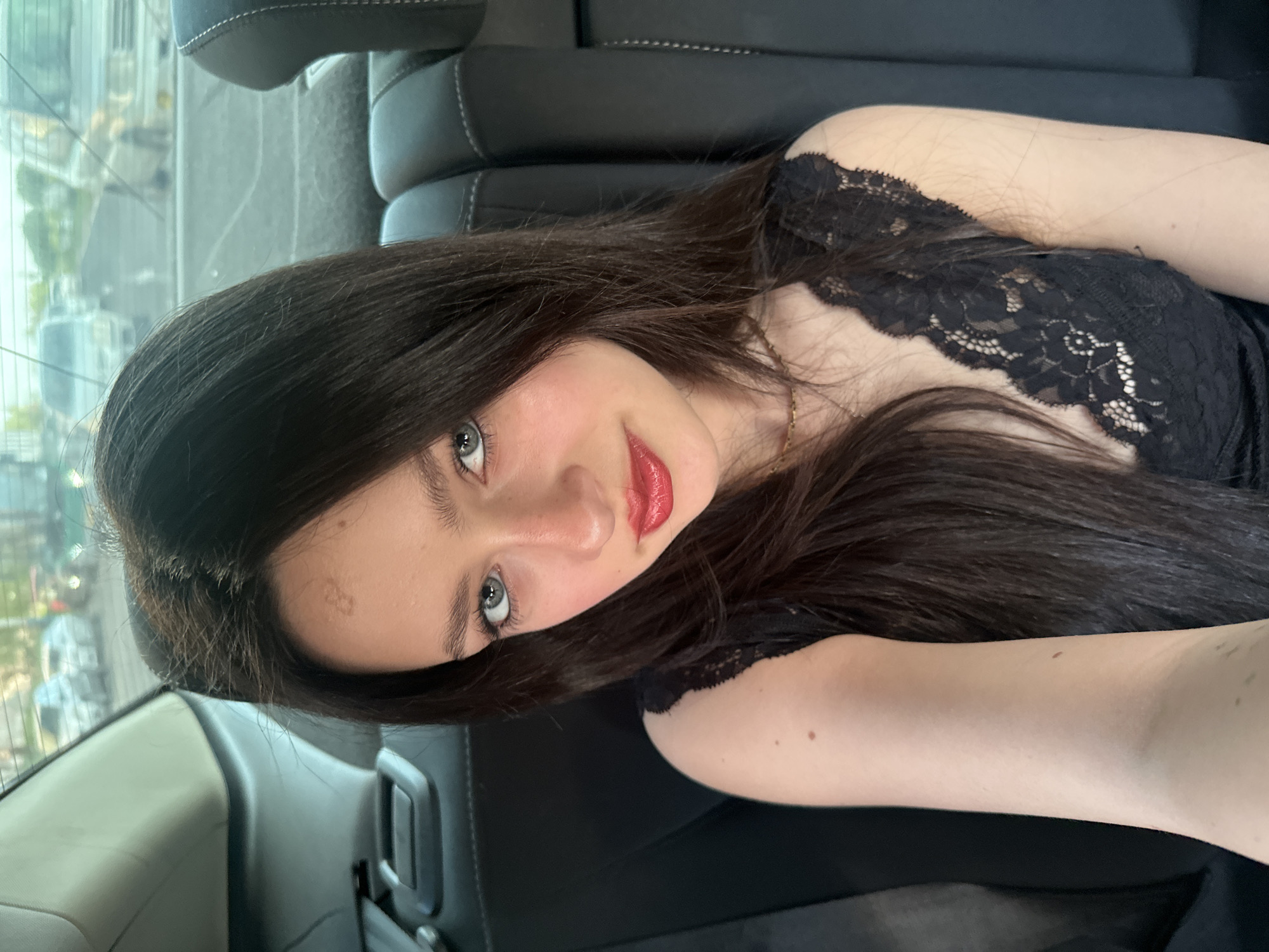 A young woman with long, dark hair, fair skin, and light-colored eyes is looking at the camera with a slight smile. She has red lipstick on. She is wearing a black, lace-trimmed top. The background suggests she is in a car. The specific location in Yerevan, Armenia, cannot be determined from the image.