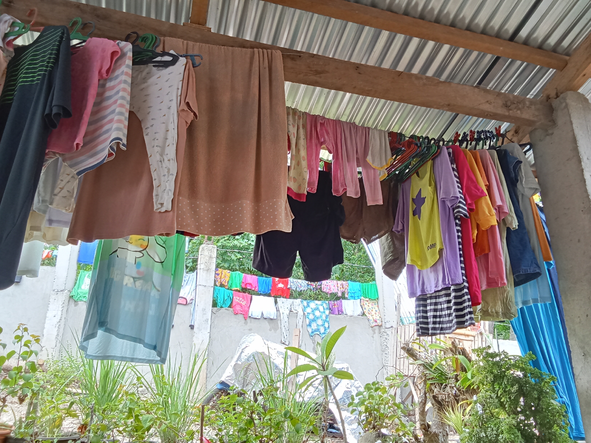 The image depicts a clothesline with various garments drying under a corrugated metal roof supported by wooden beams and a concrete pillar, likely in an outdoor or semi-outdoor area of a residential property. The clothes are hung on hangers and directly on the line, showcasing a mix of colors and patterns including t-shirts, shorts, and towels. Some clothes are vibrant, such as a yellow t-shirt with a purple star, while others are more muted. In the background, a concrete wall is visible, with more clothes hanging on a line further back. Lush green foliage and plants are present in the foreground and surrounding areas, suggesting a tropical environment. The overall scene evokes a sense of domesticity and everyday life. The location is identified as Samal City, Philippines, which is known for its tropical climate, consistent with the presence of abundant greenery and the method of drying clothes outdoors.