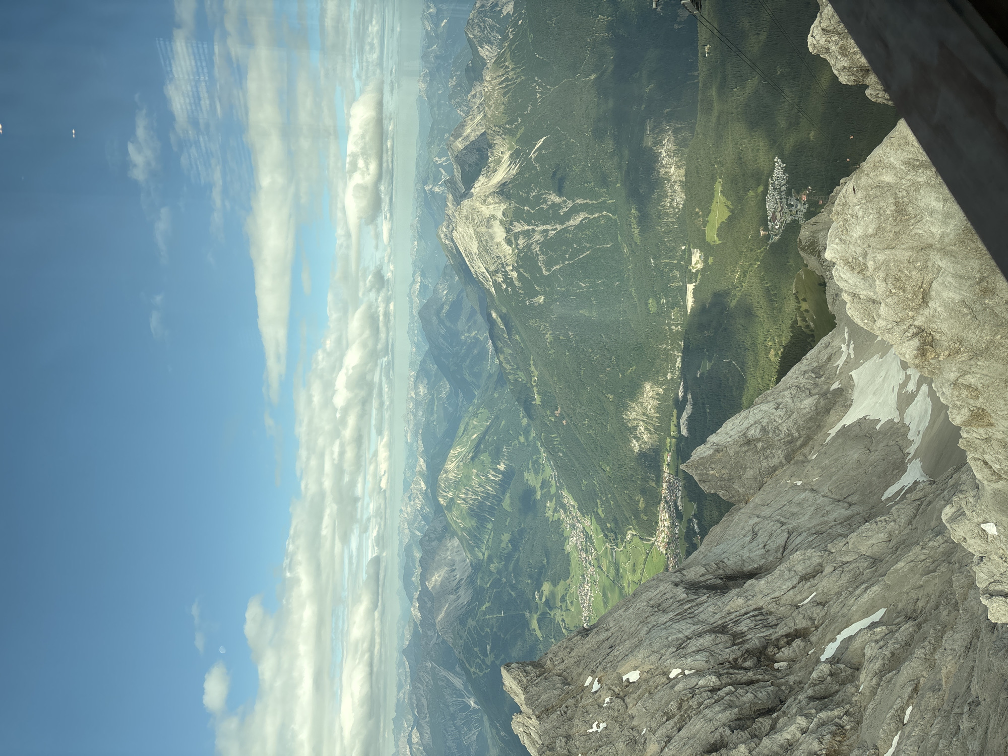 The image captures a panoramic view of the Tyrolean Alps, with a focus on the Zugspitze region near Ehrwald, Austria. In the foreground, rugged, rocky mountain peaks, partially covered with snow, dominate the left side of the frame. These peaks exhibit a rough, weathered texture.
Below these imposing mountains, a verdant valley unfolds, featuring lush green slopes and dense forests. A small town, identifiable by its buildings and infrastructure, is nestled within the valley, with roads winding through it. The landscape recedes into the distance, revealing a series of layered mountain ranges under a sky filled with scattered, fluffy white clouds against a bright blue backdrop.
The perspective suggests the image was taken from a high vantage point, possibly from a cable car or a viewing platform. A glimpse of a wooden railing or structure is visible in the bottom right corner, further supporting this idea. The overall scene is one of natural grandeur, showcasing the dramatic topography of the Alps and the charming human settlement within it.