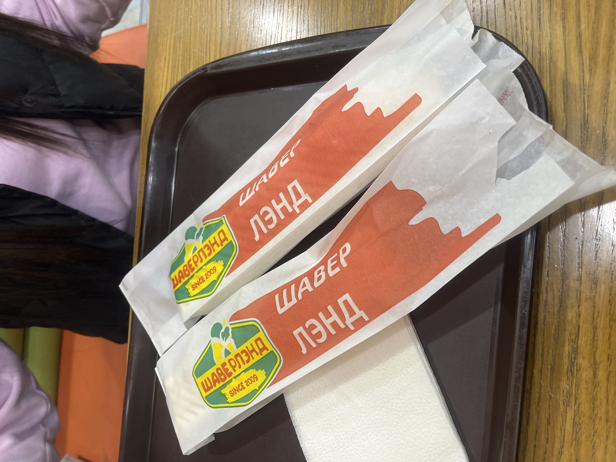 The image depicts two shawarma wraps from "Shaverland" located in Saint Petersburg, Russia, placed on a dark brown tray. The wraps are in white paper packaging with an orange design and the restaurant's logo, which includes the text "ШАВЕРЛЭНД" and "SINCE 2009." A stack of white napkins is visible between the shawarmas. The tray is set on a wooden table. In the background, a person is partially visible, wearing a light purple shirt and a black jacket. The background is a blurred mix of light colors, including orange and green.