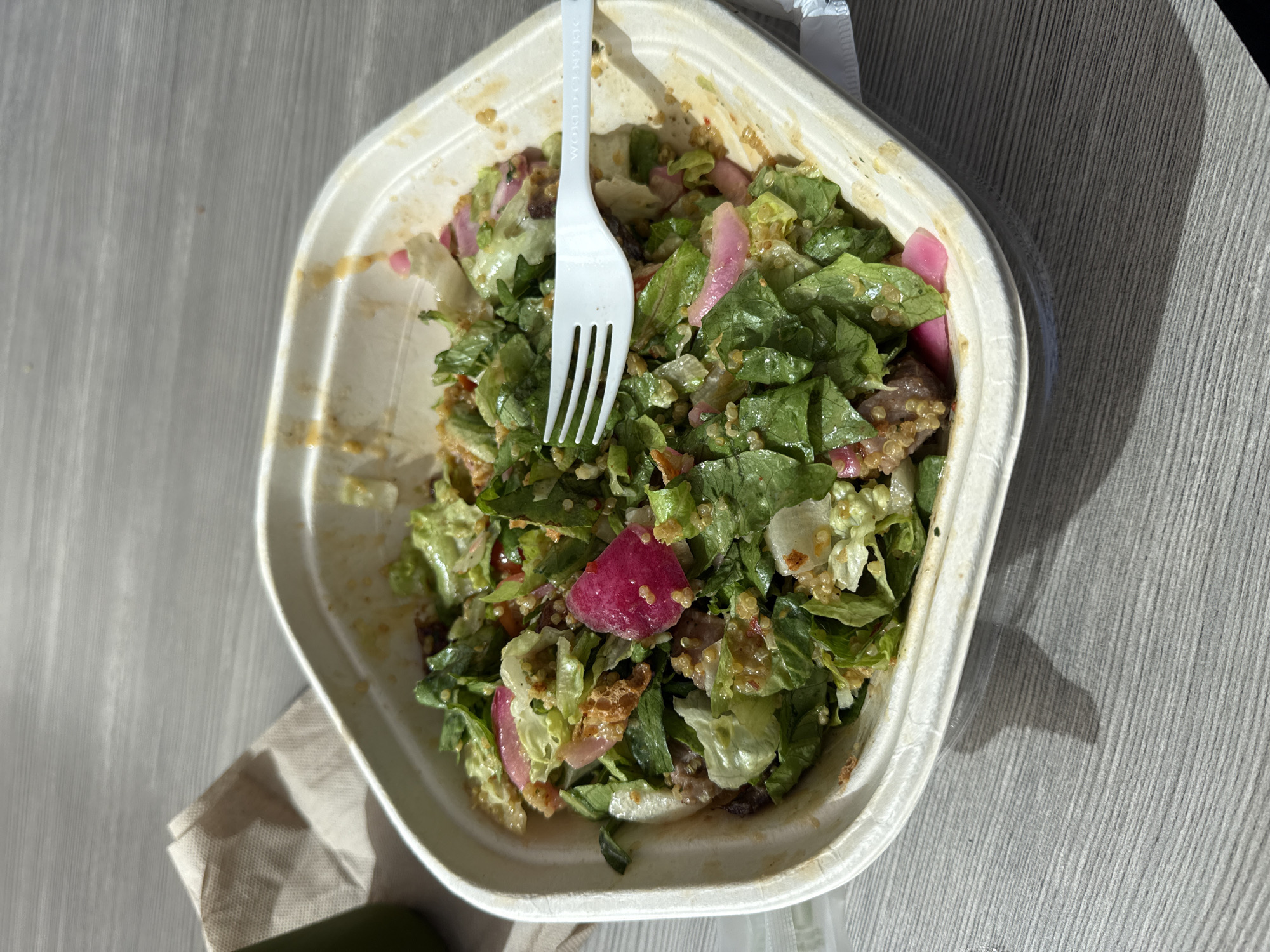 The image presents an overhead view of a fresh salad contained within a rectangular, light-colored, disposable takeout bowl. A white plastic fork, partially submerged in the salad, features the embossed text "WORLD CLASSIC" on its handle.
The salad consists primarily of chopped green lettuce, interspersed with vibrant reddish-pink radish slices and small, pearl-like grains of quinoa. A light dressing coats the ingredients, and hints of other components, possibly meat or other vegetables, are visible beneath the top layer of greens.
The takeout container is placed on a textured, light-grey surface, which appears to be an outdoor table, characterized by subtle parallel grooves. Bright natural sunlight illuminates the scene, casting sharp, dark shadows on the table surface, particularly towards the right side of the image, indicating a sunny daytime setting, likely midday or early afternoon. A crumpled brown paper napkin is partially visible beneath the container on the lower left. The overall scene suggests a casual, outdoor meal. No people are visible in the frame.