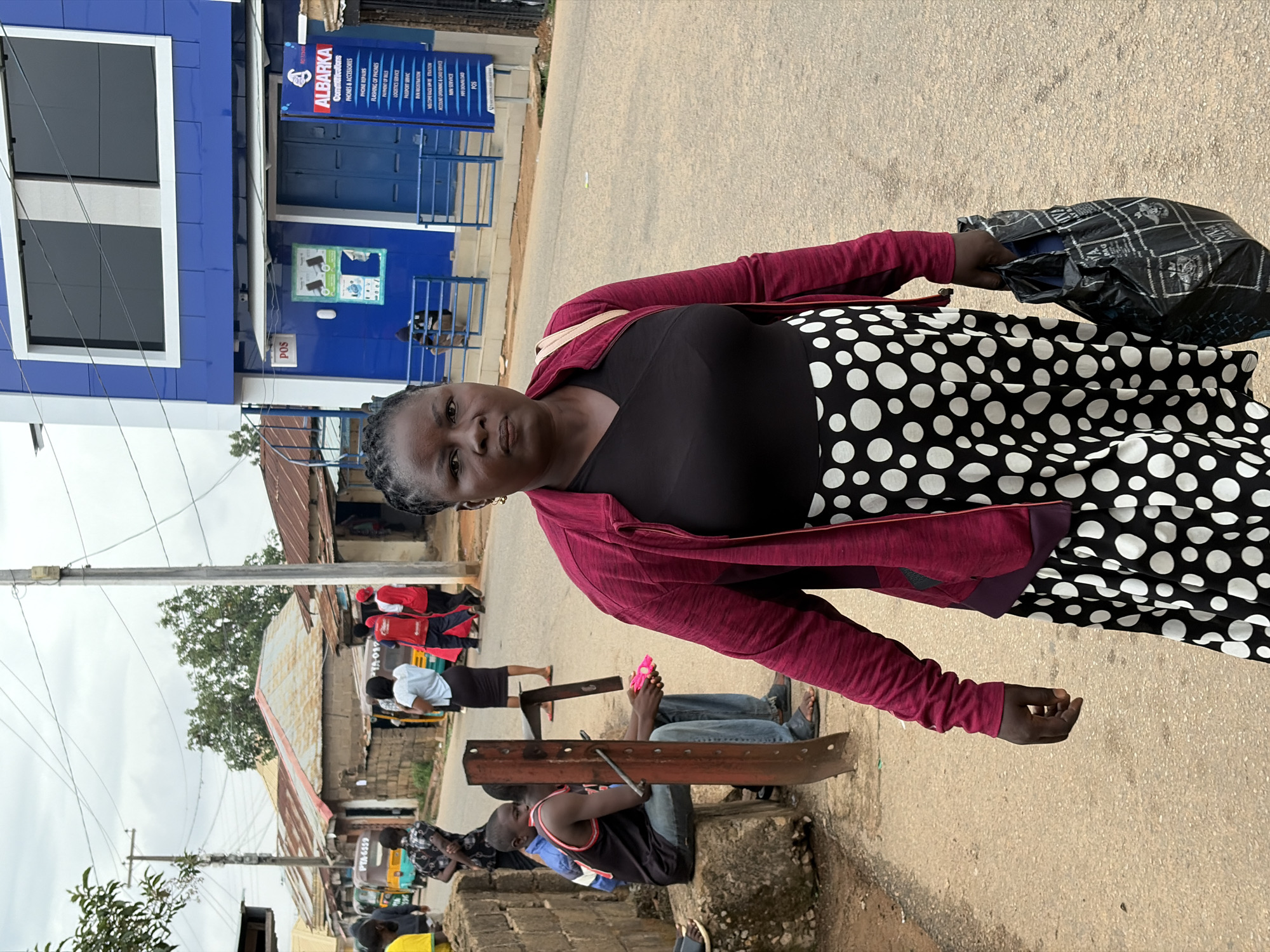 The image shows a woman standing on a street in Jos, Nigeria. She is wearing a black top, a skirt with white polka dots, and a red jacket. She is carrying a black plastic bag in her right hand. Behind her is a blue building with the name "ALBARKA Communications" printed on it. The building also lists services offered, including phone repairs, logistics service, and passport service. Further down the street, there are several people and vehicles. Some people are sitting on a concrete structure, and others are walking. There are buildings with corrugated metal roofs in the background. The street appears to be paved.