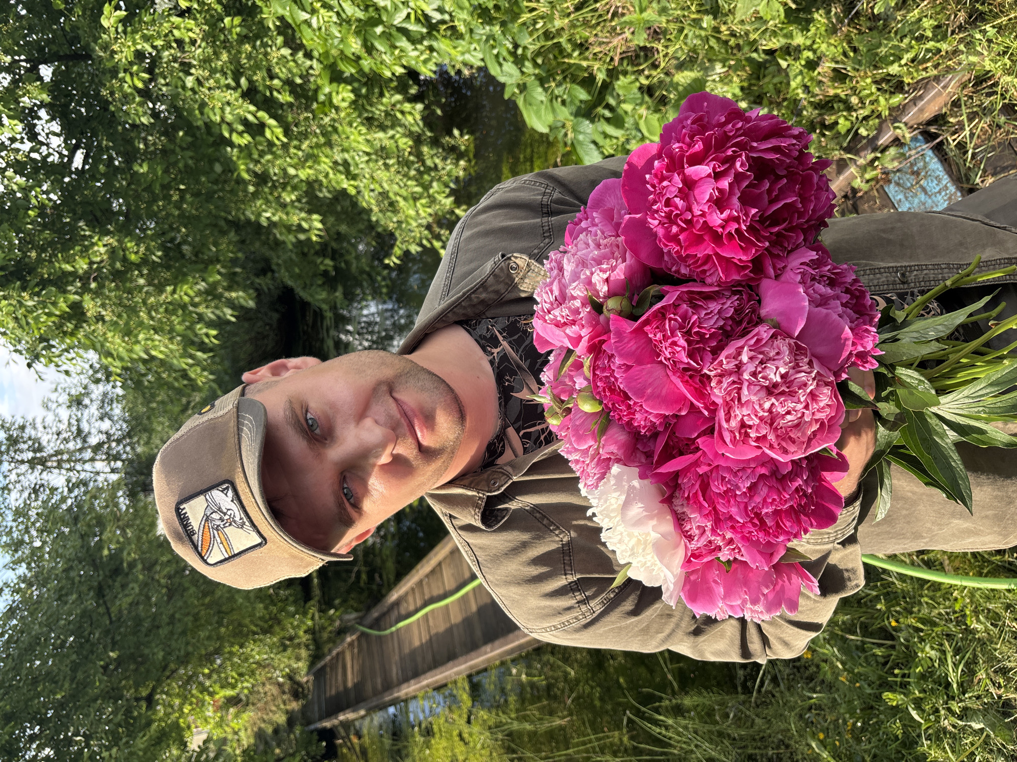 In an outdoor setting near what appears to be a small body of water and a wooden walkway, a man holds a large bouquet of pink and white peonies. He is wearing a khaki-colored button-up shirt over a dark t-shirt and a tan baseball cap with a patch depicting a cartoon rabbit. The background consists of lush green foliage and trees. The lighting suggests it is daytime, possibly late afternoon due to the warm tones. The location is identified as the city of Niva, Russia.