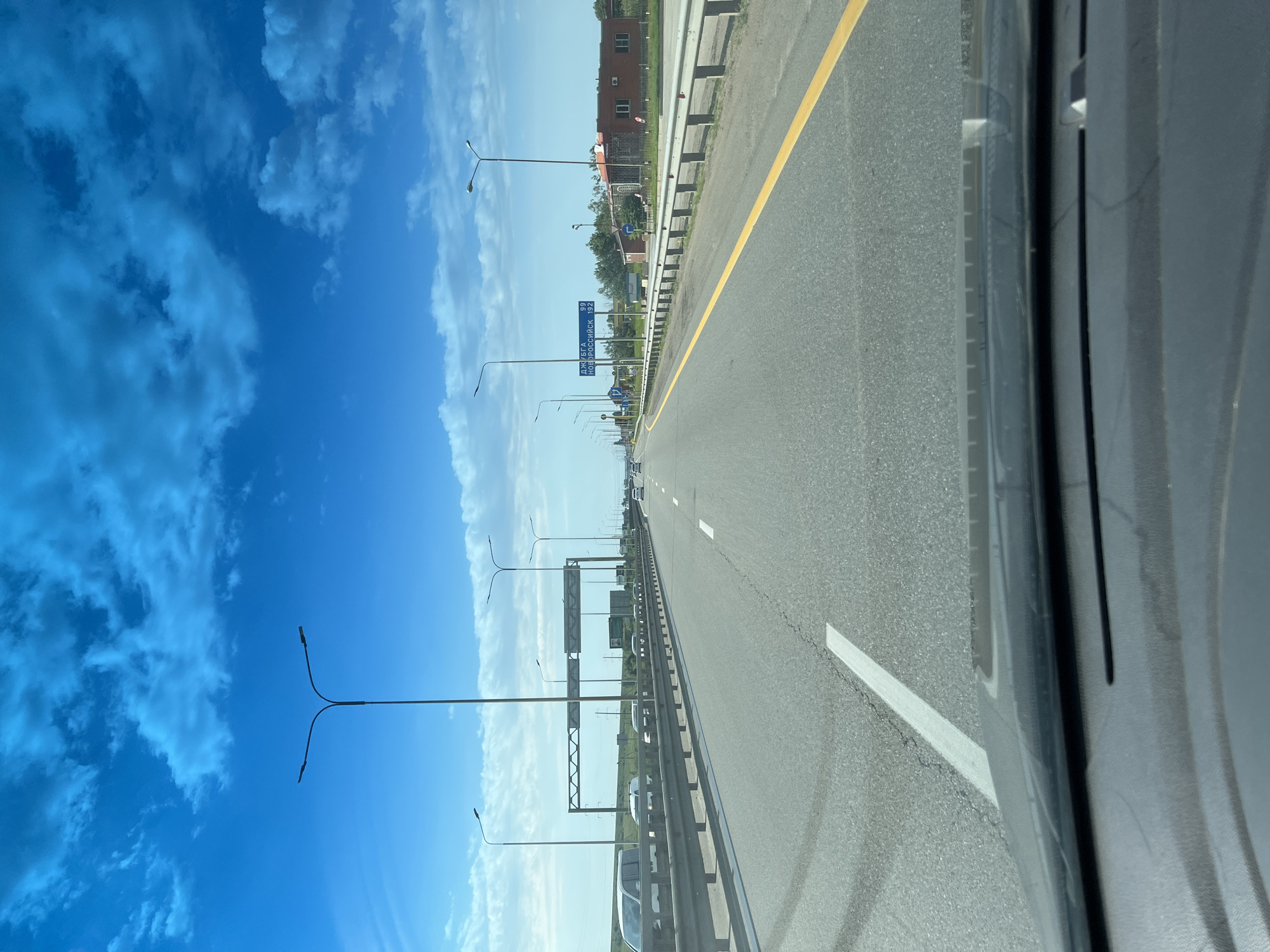 The image shows a road in what appears to be Tugurgoy, Russia, as indicated by the road sign. The road is a highway with multiple lanes, marked with yellow and white lines. There are street lights along the side of the road and guardrails. A blue sky with scattered clouds is visible above. In the distance, there is a building with windows. The image appears to be taken from inside a vehicle.