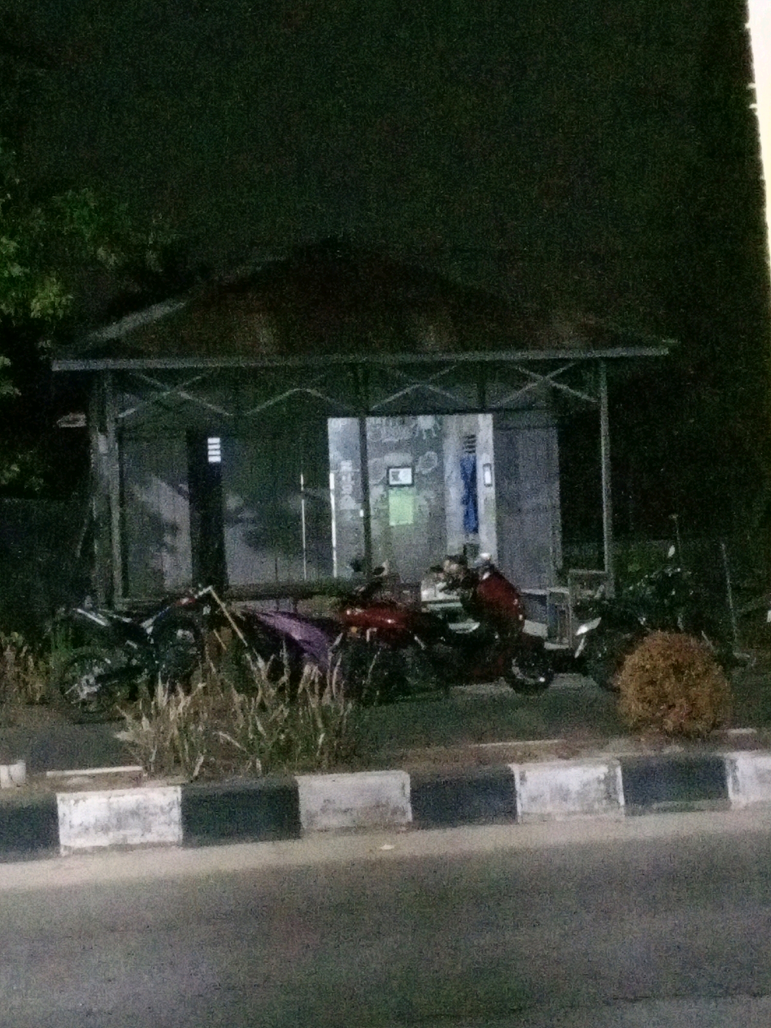 The image shows a nighttime scene in front of a small, open-sided shelter with a sloped roof. Inside the shelter, there are some illuminated panels or signs. Several motorcycles are parked outside the shelter, with some partially obscured by vegetation. A street with a curb is visible in the foreground. The overall setting suggests an outdoor area, possibly a bus stop or a rest area, in a dimly lit environment. There are no people visible in the image.