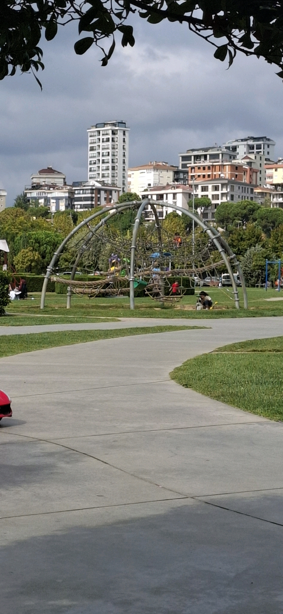 The image shows an outdoor children's playground in Maltepe, Turkey, on an overcast day. The playground features a large, geodesic dome climbing structure made of metal bars and ropes, with various platforms and climbing elements. In the background, several multi-story apartment buildings and trees are visible. A paved pathway curves through the grassy area in the foreground. There are a few people scattered throughout the scene; one person is sitting on the grass near a red object on the left side of the pathway, and another person is playing in a sandbox in the middle ground to the right of the climbing structure. Children are also visible on the climbing structure. The sky is cloudy.