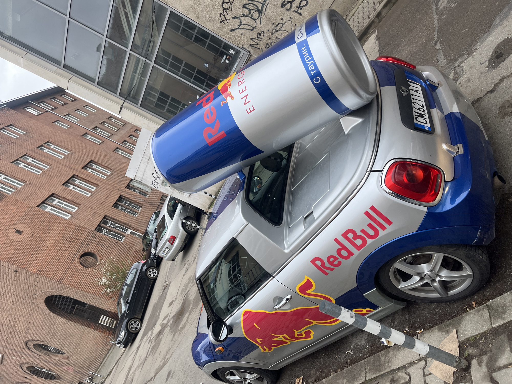 The image captures a promotional vehicle parked on a city street in Sofia, Bulgaria. The car is a silver and blue Mini Cooper, customized with Red Bull branding, including a large Red Bull can mounted on its roof. The license plate is visible and reads "CM.6211.AJ" with "BG" for Bulgaria. The car is parked beside a black and white bollard on a street with visible pavement. In the background, there's a multi-story brick building with numerous windows, along with a more modern structure featuring glass panels above. Two other cars, one silver and one black, are parked further down the street. Graffiti is present on the wall of the modern structure above the car. The environment appears to be urban, with a mix of older and more contemporary architecture.