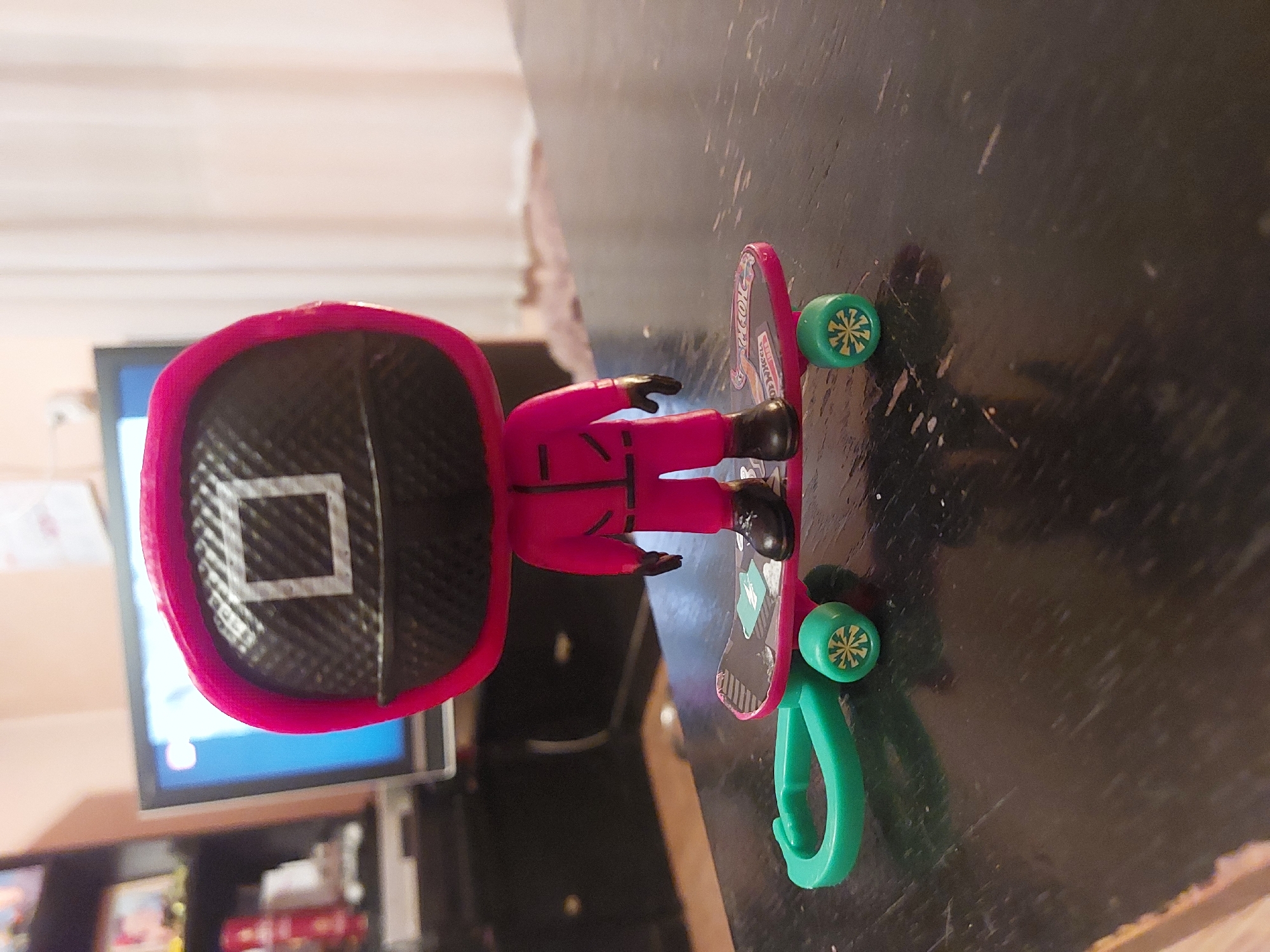A close-up shows a toy figurine of a character from the Netflix series "Squid Game" standing on a pink toy skateboard on a dark wooden surface. The figurine is wearing a pink jumpsuit and a black helmet with a white square on the front. The skateboard has green wheels and a green plastic clip attached to one end. In the blurred background, a television screen is visible, along with a dark cabinet and various items on a shelf. There is no information suggesting the photograph was taken in Tsarevo, Bulgaria.