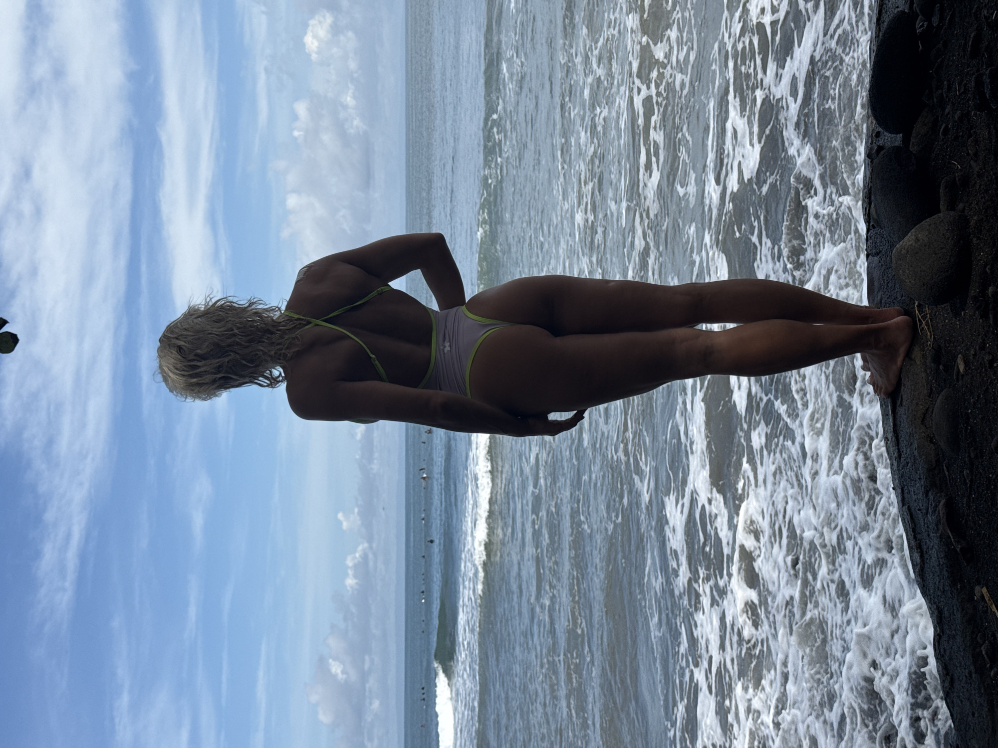 A woman stands on a rocky outcrop next to the ocean, facing the water with her back to the camera. She wears a pale bathing suit with neon green trim. Her blond hair is loose and curly. The sky is blue with scattered clouds. Waves crash against the rocks. The location is the city of Unknown, Unknown.