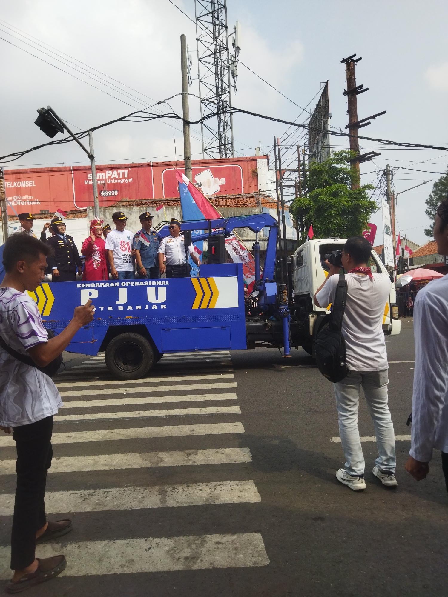 In Banjar, Indonesia, a man in the foreground, wearing a white t-shirt with purple stripes and black pants, stands on a pedestrian crossing, looking at his phone. He is positioned to the left of a blue truck with yellow markings and the text "KENDARAAN OPERASIONAL PJU" and "TA BANJAR" printed on its side. Several people, including men in police or formal uniforms and a woman in traditional attire, are standing on the back of the truck, some holding flags. To the right, a man with a red bandana around his neck is taking a photograph of the truck with a DSLR camera. Another person, partially visible on the far right, also appears to be observing the scene. The truck has a crane attached. In the background, tall communication towers and a large billboard are visible, along with some trees and buildings. The sky is overcast.