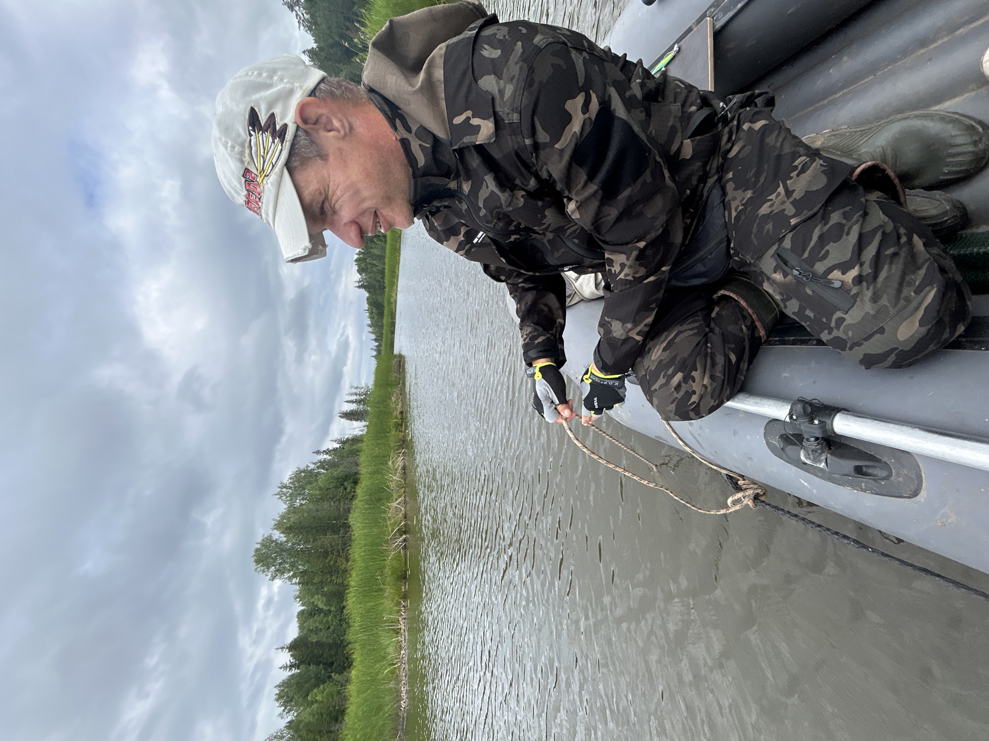 A man in a camouflage jacket and pants, a white baseball cap, and green rubber boots is sitting in a gray inflatable boat on a body of water. He is holding a rope in his hands, and the rope is tied to the boat. The man is looking to his right and smiling. The boat is surrounded by green vegetation and trees on the bank of the water. The sky is overcast. The scene appears to be in a rural or natural setting, possibly near Detskii Dom (Bashenino), Russia, given the natural landscape.