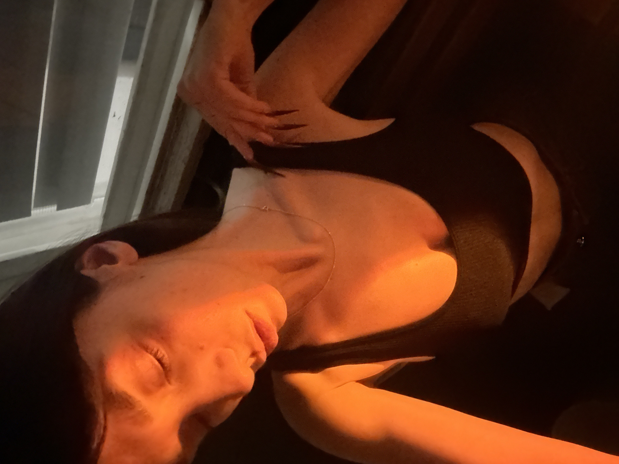 A woman with long, dark hair is lying down, eyes closed. She is wearing a dark top and a delicate necklace. Her hand is near her chest, with long, dark nails visible. Light is shining on her face, creating a warm, orange glow. Blinds are partially visible in the background. The location is unknown.