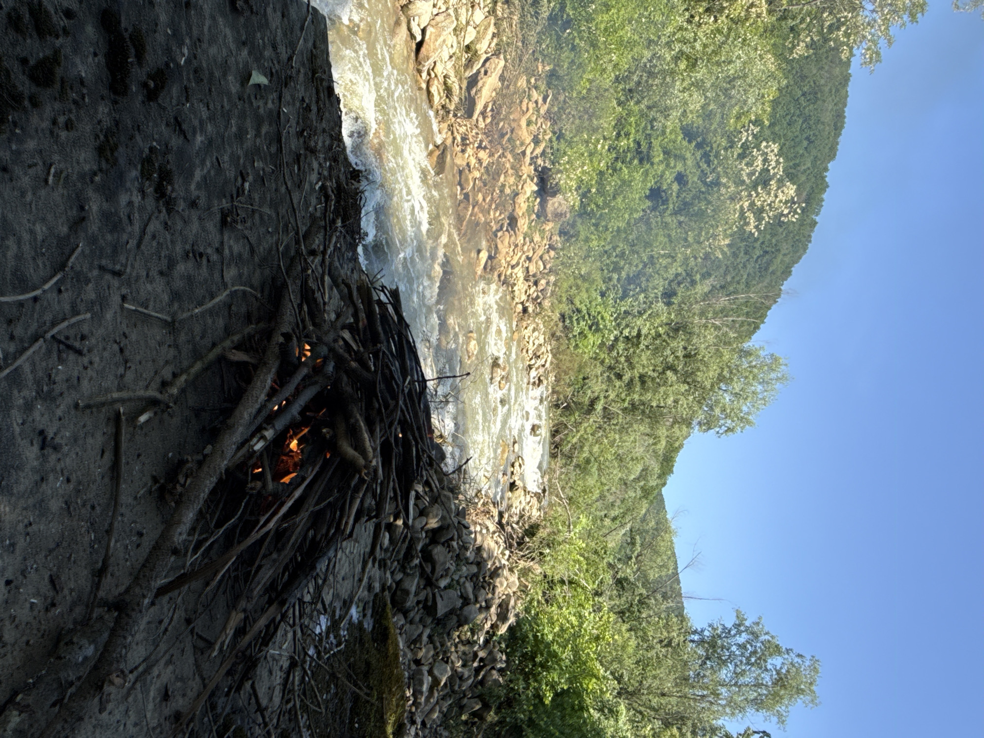 A landscape shot, rotated ninety degrees, features a bonfire constructed of sticks near a rushing river. The river, with its turbulent, brownish water, flows from the upper left to the lower right. Adjacent to the fire is a mix of dirt and small rocks. Across the river, a hillside is densely covered with green trees and other vegetation. Above the hillside is a clear, blue sky. The location is Stara Rechka, Bulgaria.