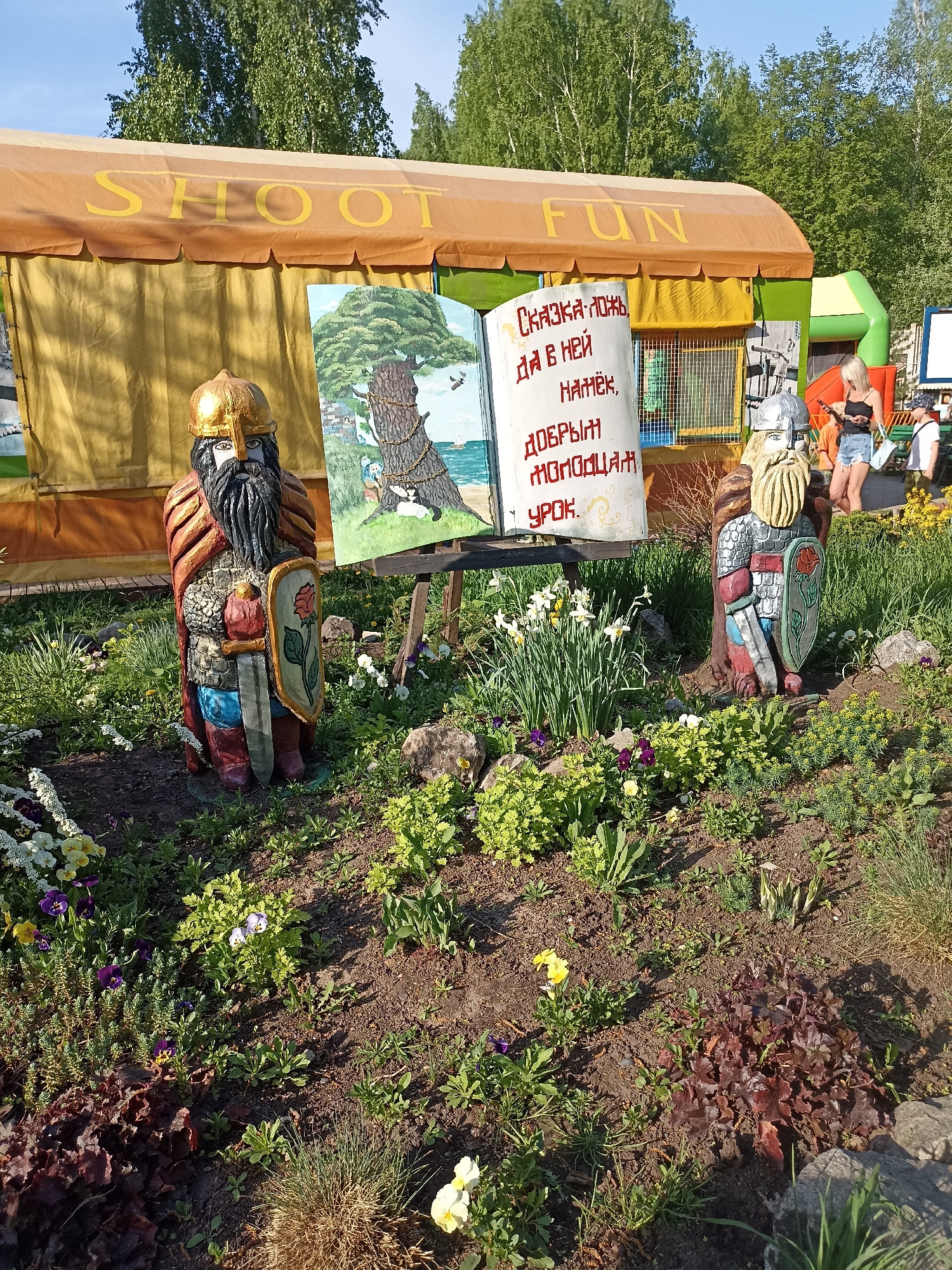 The scene captures a small, decorative garden display in what appears to be a public area. Two carved wooden statues of what resemble Viking figures are placed in the garden. The statue on the left has a gold helmet and beard, while the one on the right has a silver helmet. Each holds a shield with a rose design and a sword.
Between the statues is an open book displayed on an easel. The left page shows a painted scene of a tree by the water. The right page contains text in Russian, written in red ink.
The garden itself features a variety of flowers and plants, including white daffodils, pansies, and other greenery.
In the background, a portable structure with "SHOOT FUN" written on it is visible, suggesting an amusement area. People can be seen in the background, including a woman in denim shorts looking at her phone. The overall environment indicates a recreational or tourist area.