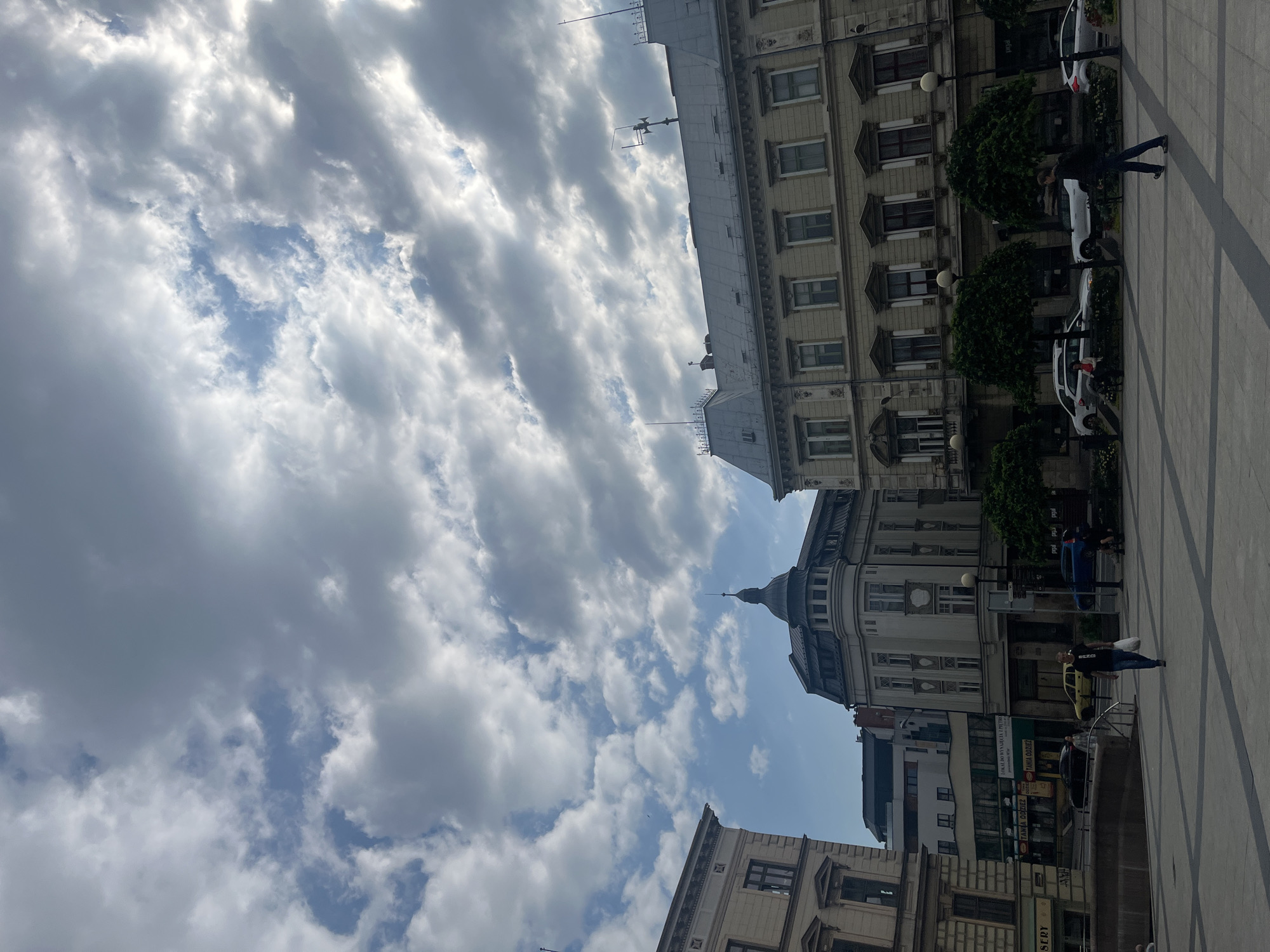 An outdoor, eye-level, medium shot captures a city square in the Unknown region of Unknown. The scene is dominated by a dramatic sky filled with a mix of bright white and dark gray clouds, with patches of blue sky visible. Several buildings with ornate architecture line the square. On the left, a light-colored building with multiple windows and decorative elements is partially visible. In the center, a lighter colored building with a domed roof and a spire stands out. To the right, a larger, light-yellow building features a long facade with numerous windows and decorative detailing, topped with a grey roof with several antennae.
The foreground shows a paved square with a geometric pattern. A few trees with dense, rounded canopies are planted along the edge of the square in front of the buildings. Several people are present, including a man walking towards the right side of the frame, carrying a white bag, and another person walking away from the viewer on the right. Cars are parked and driving in the background. A street lamp is visible in front of the light-yellow building. The lighting suggests it is daytime, with sunlight filtering through the clouds, creating variations in light and shadow.