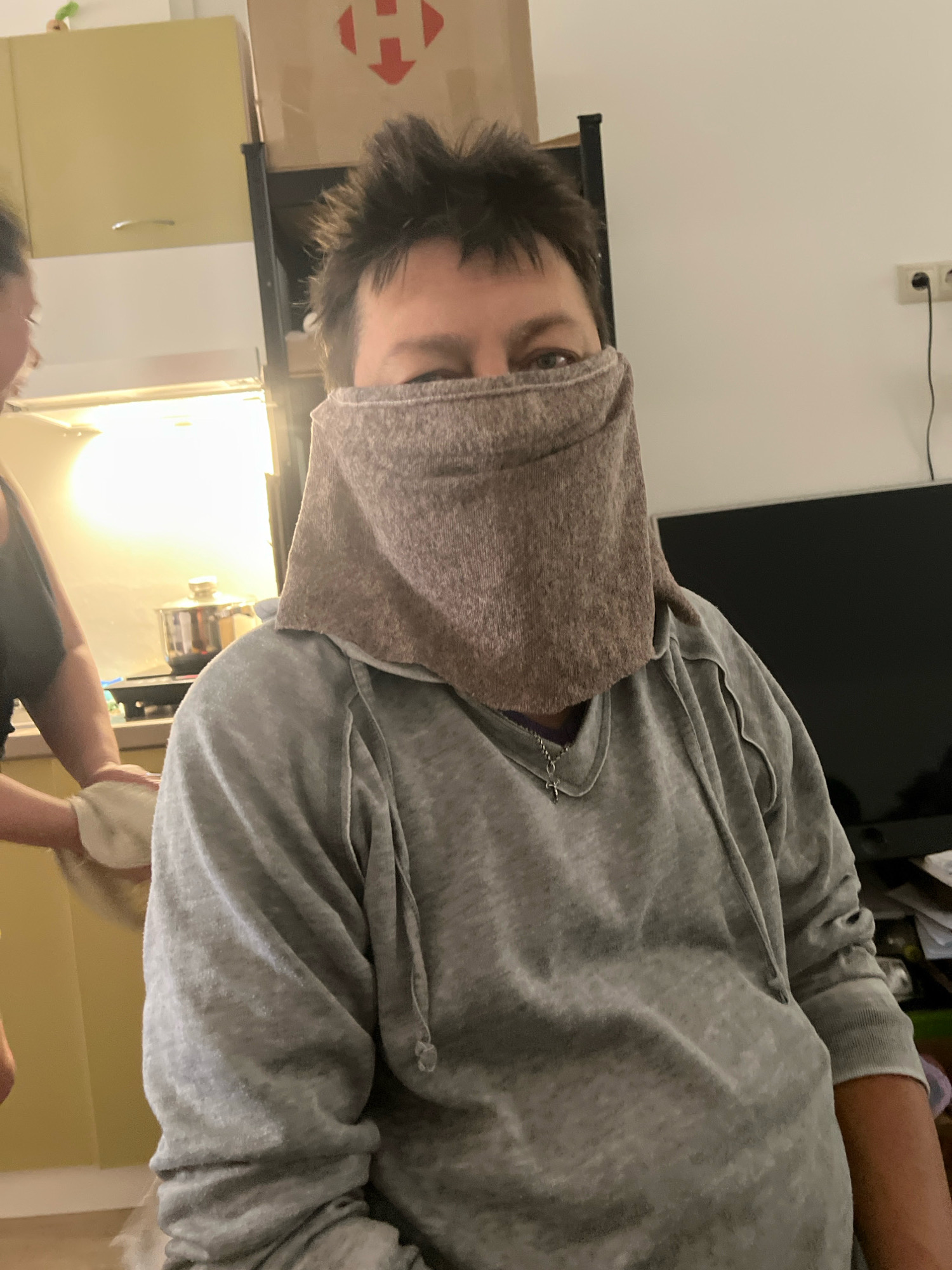 The image shows a person in the foreground wearing a gray hoodie and a brown cloth covering their face, with only their eyes visible. The person is indoors, in what appears to be a kitchen or living area. In the background, there are kitchen cabinets, a shelf with a box, and a television. Another person is partially visible on the left, wearing a dark top and holding a white cloth. The overall setting suggests a domestic environment. The image does not contain any information that would indicate it was taken in the city of Truskavets, Ukraine.