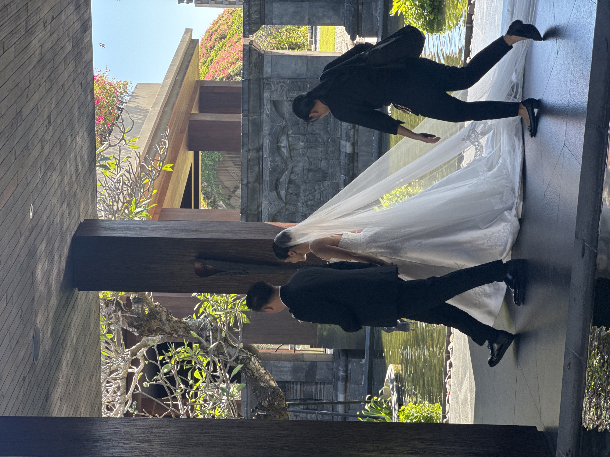 The image captures a wedding scene in Kuta Selatan, Indonesia. A bride in a white wedding gown and veil stands with a groom in a black suit. A second man, also in a black suit, stands beside the bride. The setting appears to be an outdoor space near a body of water. The architecture includes stone structures, and there is greenery including trees and plants.
