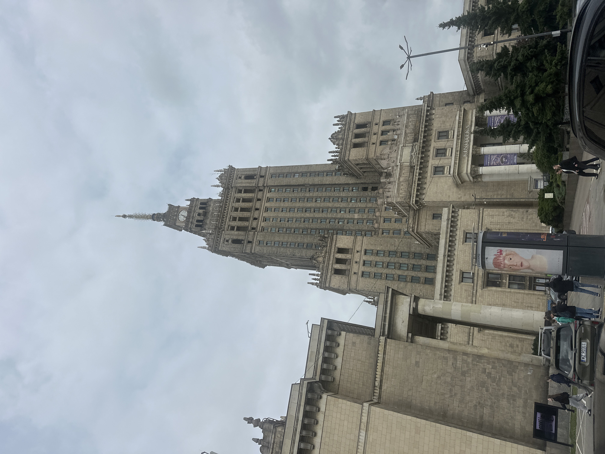 The image shows the Palace of Culture and Science in Warsaw, Poland, a prominent landmark in the city. The building is a massive, Stalinist-era skyscraper with a distinctive clock tower. In front of the building, there are several cars, including a dark-colored SUV with a Polish license plate. People are walking on the sidewalk and near the cars, some appearing to be tourists. There is also a cylindrical advertising pillar displaying a poster of a person's face. The sky is overcast, suggesting cloudy weather. The architectural style of the building is imposing and monumental, characterized by its sheer scale and ornate details. The surrounding area appears to be an urban environment with trees and streetlights. The overall impression is one of a busy public space in front of a significant historical building.