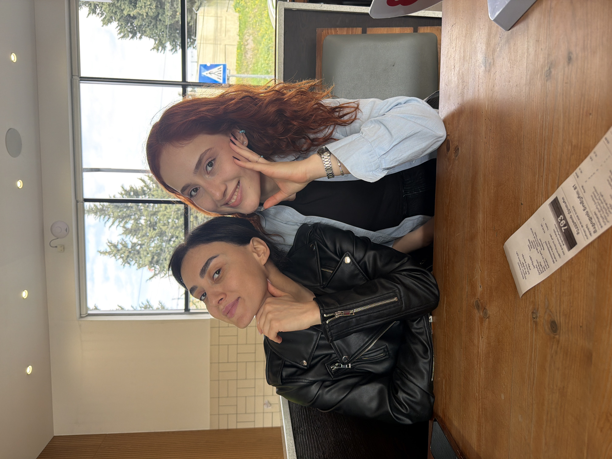A portrait-oriented photo, rotated 90 degrees clockwise, captures two women in what appears to be a casual dining setting like a Wendy's restaurant, given the visible text. The women are seated at a light brown wooden table, with large windows behind them showcasing an outdoor scene.
The woman positioned higher in the frame has long, wavy reddish-brown hair and is wearing a light blue collared shirt over a black top. She smiles warmly at the camera, with one hand raised near her cheek, showing dark nail polish and a silver watch on her wrist. She also has small turquoise earrings.
Below her, the second woman has dark hair and is wearing a black leather jacket with multiple zippers over a dark top. She offers a subtle smile, gazing towards the viewer, with one hand gently resting near her neck. Both women appear relaxed and content.
Through the windows, green trees and a clear sky with some clouds are visible, suggesting daytime. A blue and white pedestrian crossing sign can be seen outside, indicating a street or sidewalk nearby.
On the right side of the wooden table, a paper receipt is partially visible. The OCR of this receipt indicates it's from "Wendy's Varkatili" and states "შეკვეთის ქვითარი" (Order Receipt), "მაგიდის ნომერი: 81" (Table Number: 81), and a total amount of "62.90 GEL," confirming the location context of Tbilisi, Georgia. Ceiling lights and a smoke detector are also visible in the upper-left corner of the frame.