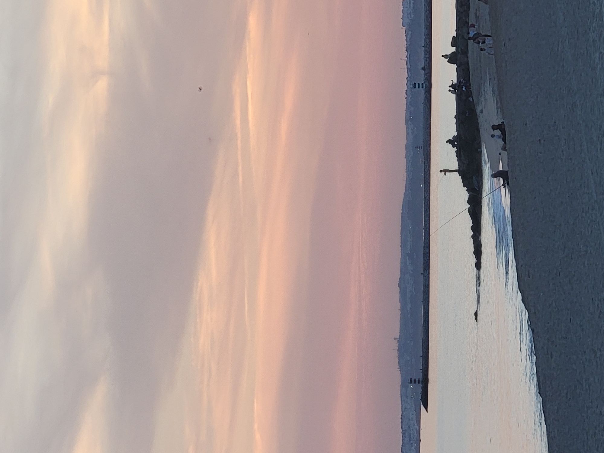 The image depicts a tranquil sunset scene at Quarteira beach in Portugal. The sky is a blend of soft pinks and purples, with wispy clouds stretching across the horizon. In the foreground, a sandy beach meets the calm, reflective water of the Atlantic Ocean. Several people are scattered along the shoreline and on a rocky breakwater. Some individuals are seated on the sand, while others are standing or walking. On the breakwater, a few figures are engaged in fishing, with fishing rods extended. A lighthouse is visible in the distance on the breakwater, and another structure, possibly another lighthouse, can be seen further along the coastline. The overall atmosphere is one of peace and relaxation, characteristic of a sunset by the sea.