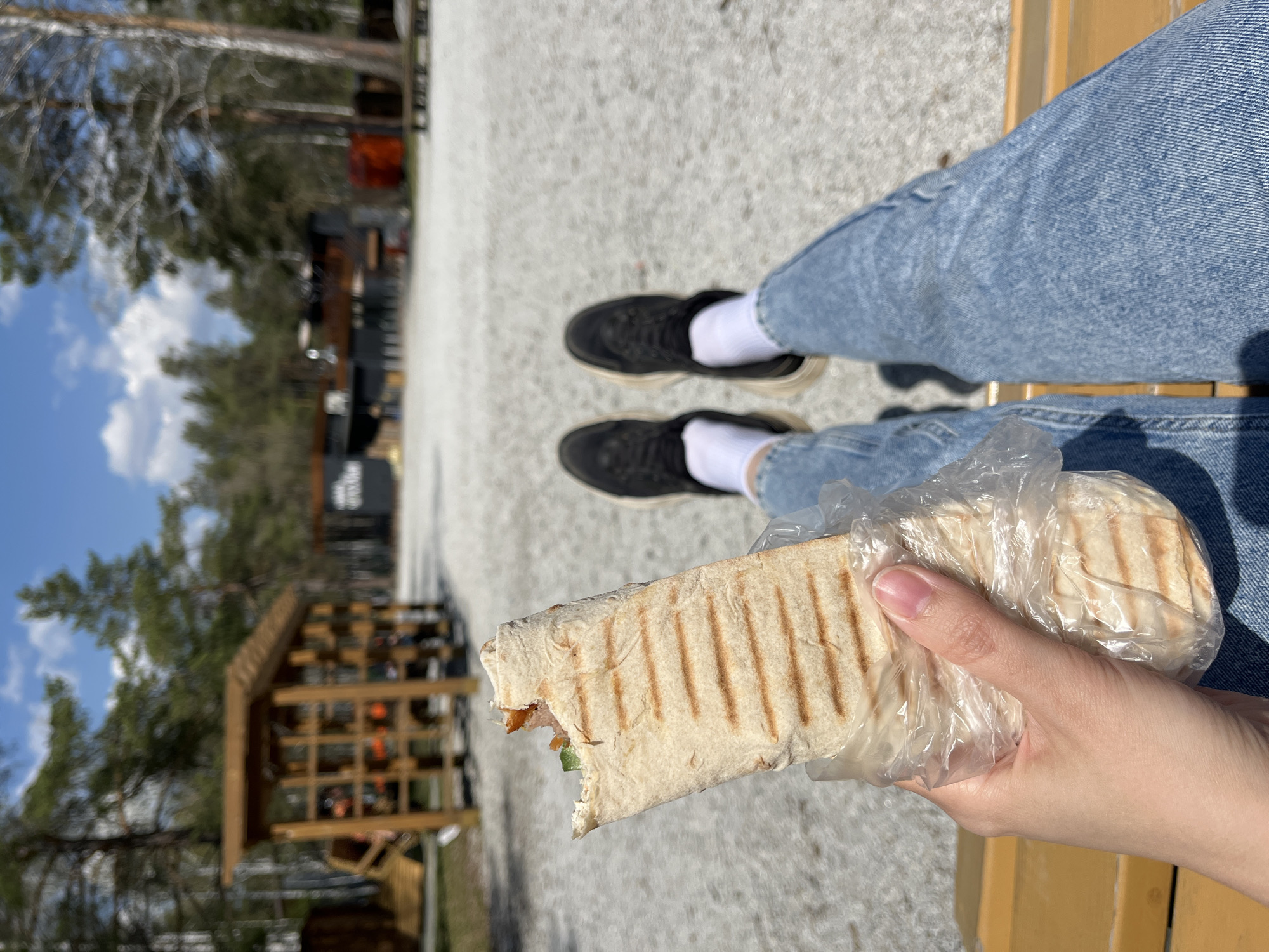 A first-person perspective image depicts a person sitting outdoors, possibly on a bench in Verkhnyaya Pyshma, Russia. The person is wearing blue jeans, white socks, and black shoes. They are holding a partially eaten shawarma in a clear plastic bag in their right hand. The shawarma has grill marks on the wrap.
The background shows a partly cloudy sky, trees, and a wooden structure with visible seating. The ground appears to be concrete. Buildings are visible in the distance.