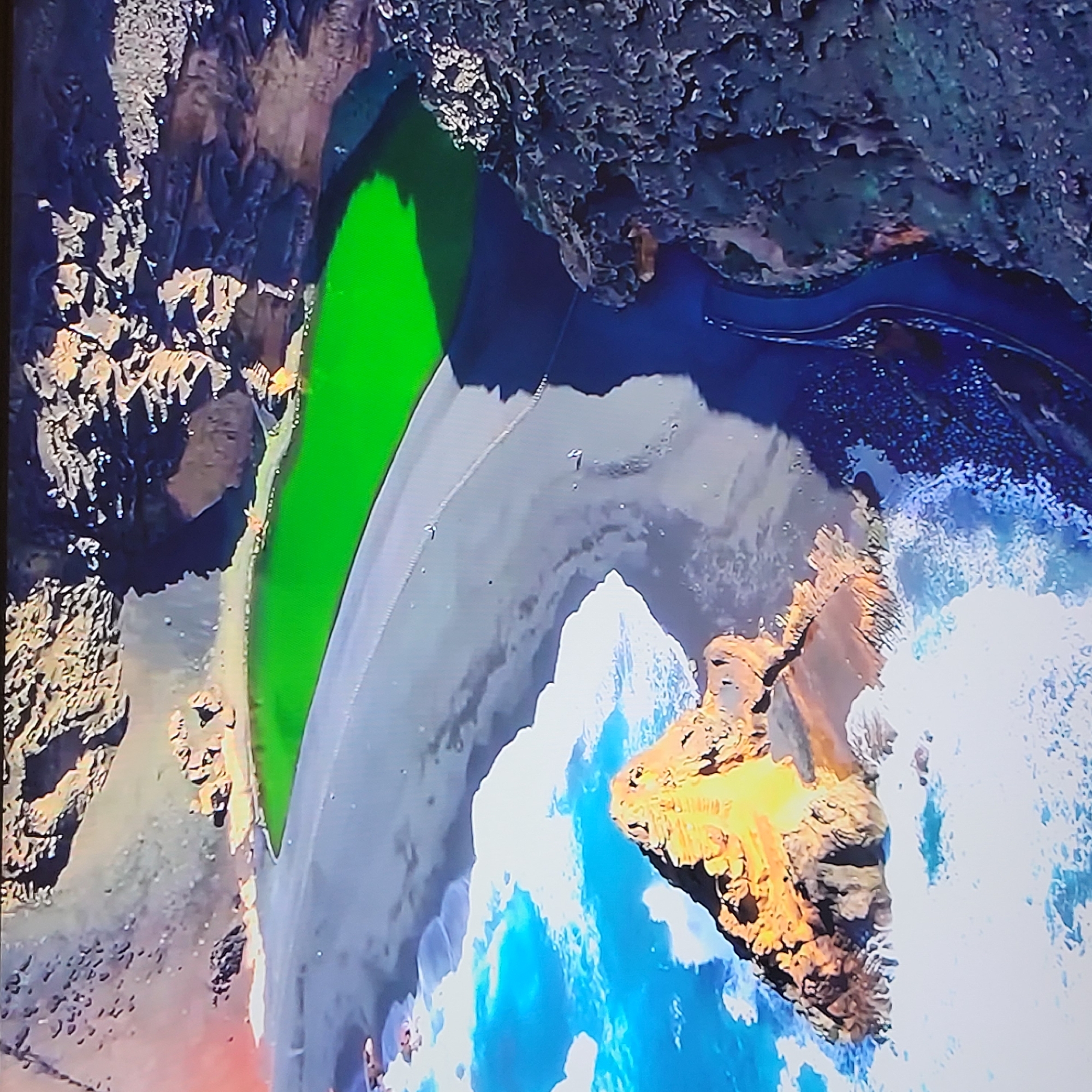 The image shows a high-angle view of a coastal landscape. A strip of black sand beach separates rocky cliffs and the ocean. Waves crash against the rocks at the lower right. A patch of bright green vegetation is visible near the left cliffs. The water is dark blue. No people are apparent. The scene does not depict Yerevan, Armenia, as it lacks any urban characteristics.